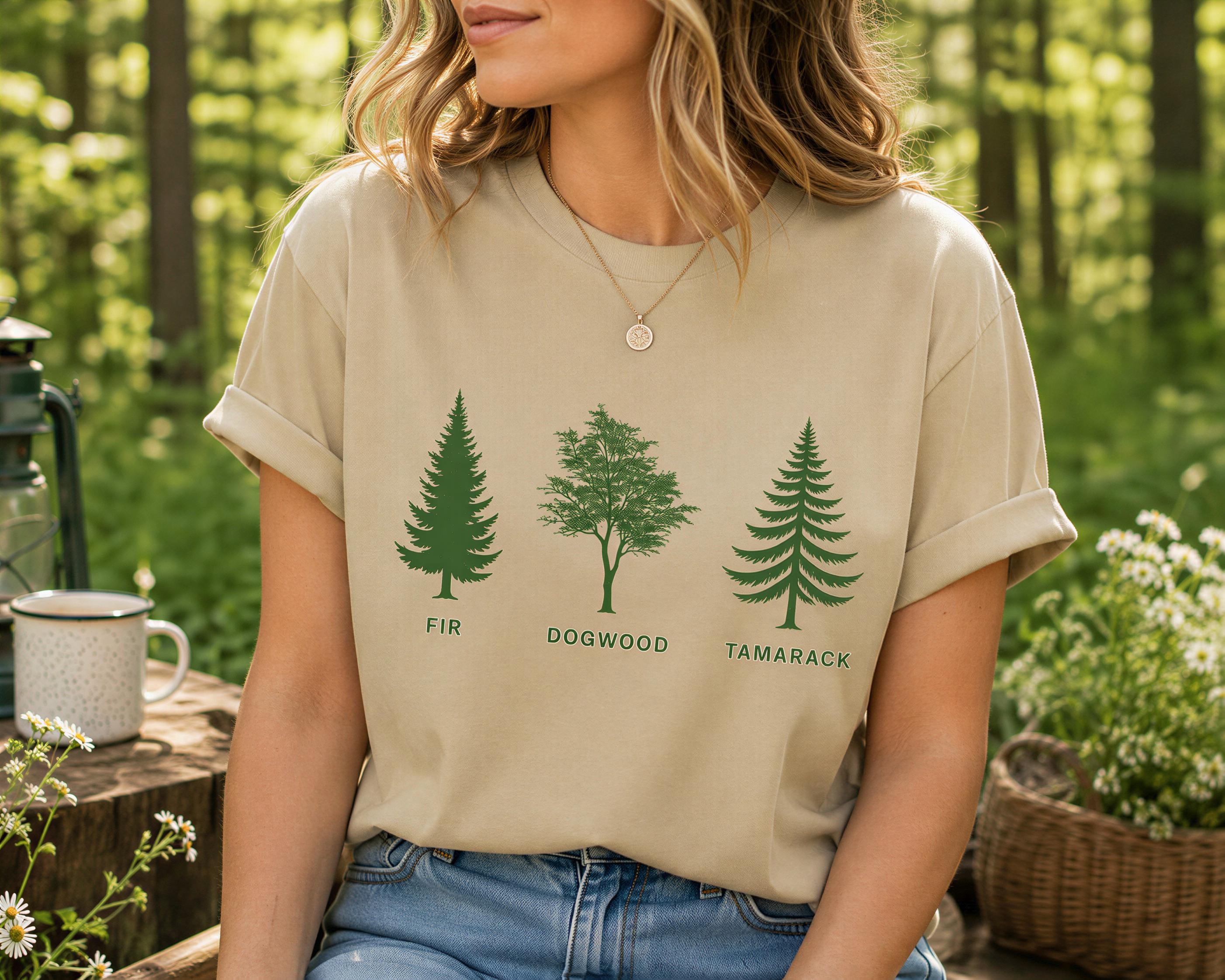 A woman wearing a beige t-shirt with three green tree silhouettes printed on it, each labeled with a different type of wood: fir, dogwood, and tamarack.