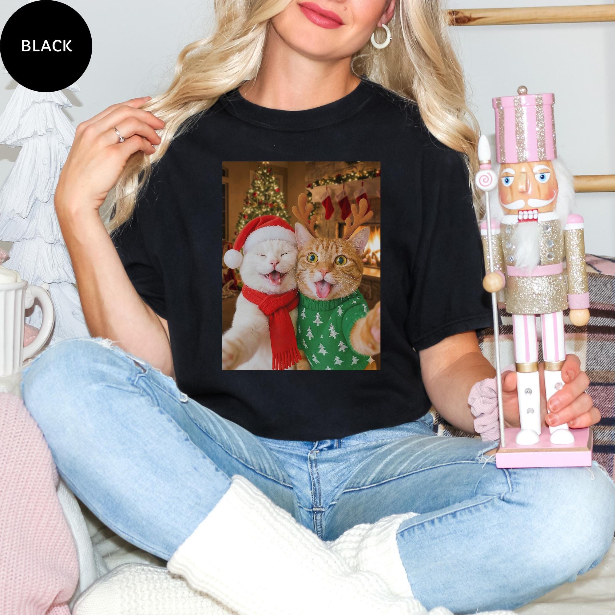 A woman is sitting on a couch, wearing a black t-shirt with a picture of two cats wearing Christmas sweaters. She is holding a nutcracker and a small Christmas tree.