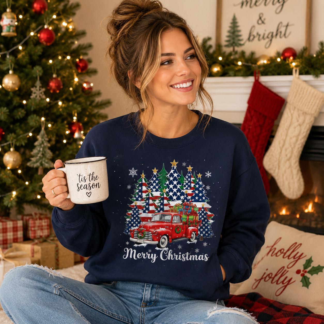 A woman is sitting in front of a Christmas tree, holding a mug with the phrase "Tis the season" written on it. She is wearing a navy blue sweatshirt with a graphic of a red truck and Christmas trees.