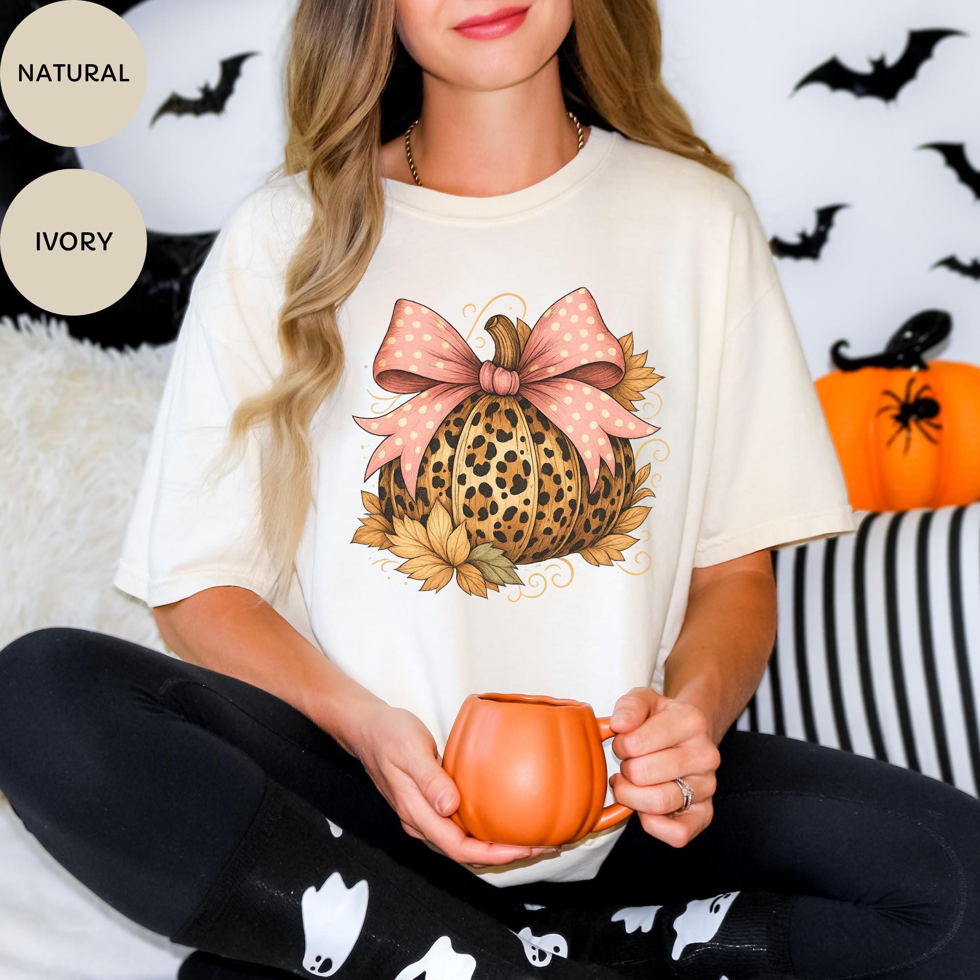 A woman wearing a white t-shirt with a pumpkin graphic and a pink bow, holding a mug, with Halloween decorations visible in the background.
