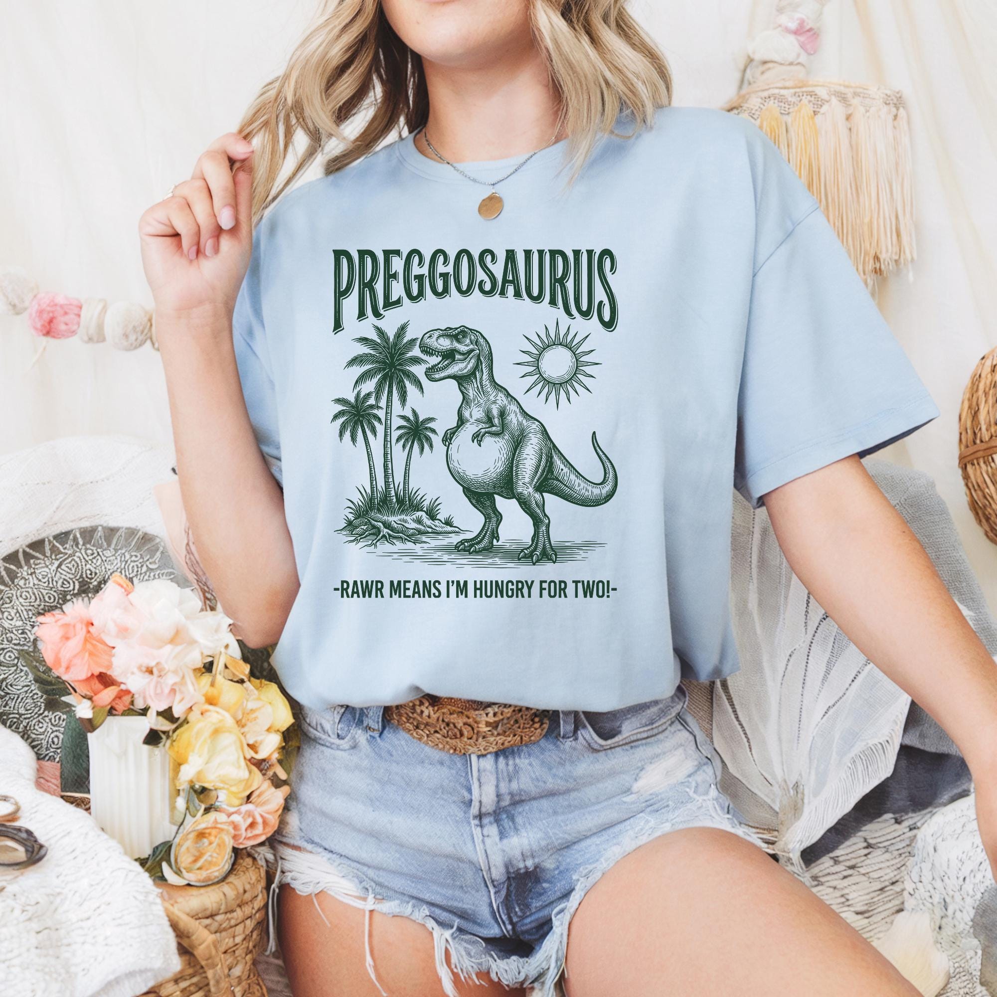 A woman wearing a light blue t-shirt with a graphic of a dinosaur and text that says "Pregosaurus" and "RAW MEANS I'M HUNGRY FOR TWO-".