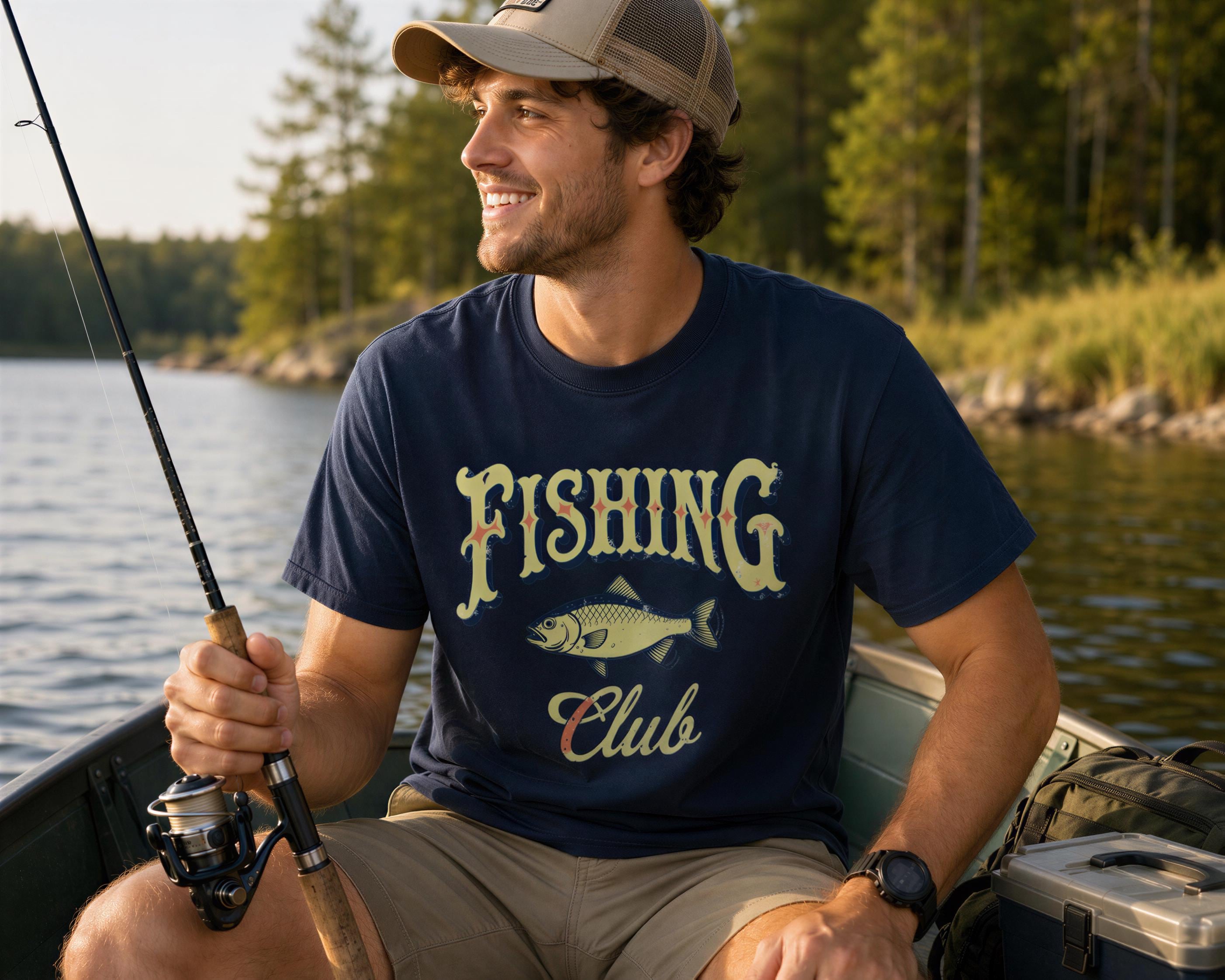 A man wearing a fishing club t-shirt and a baseball cap is sitting in a boat on a lake, smiling and holding a fishing rod.