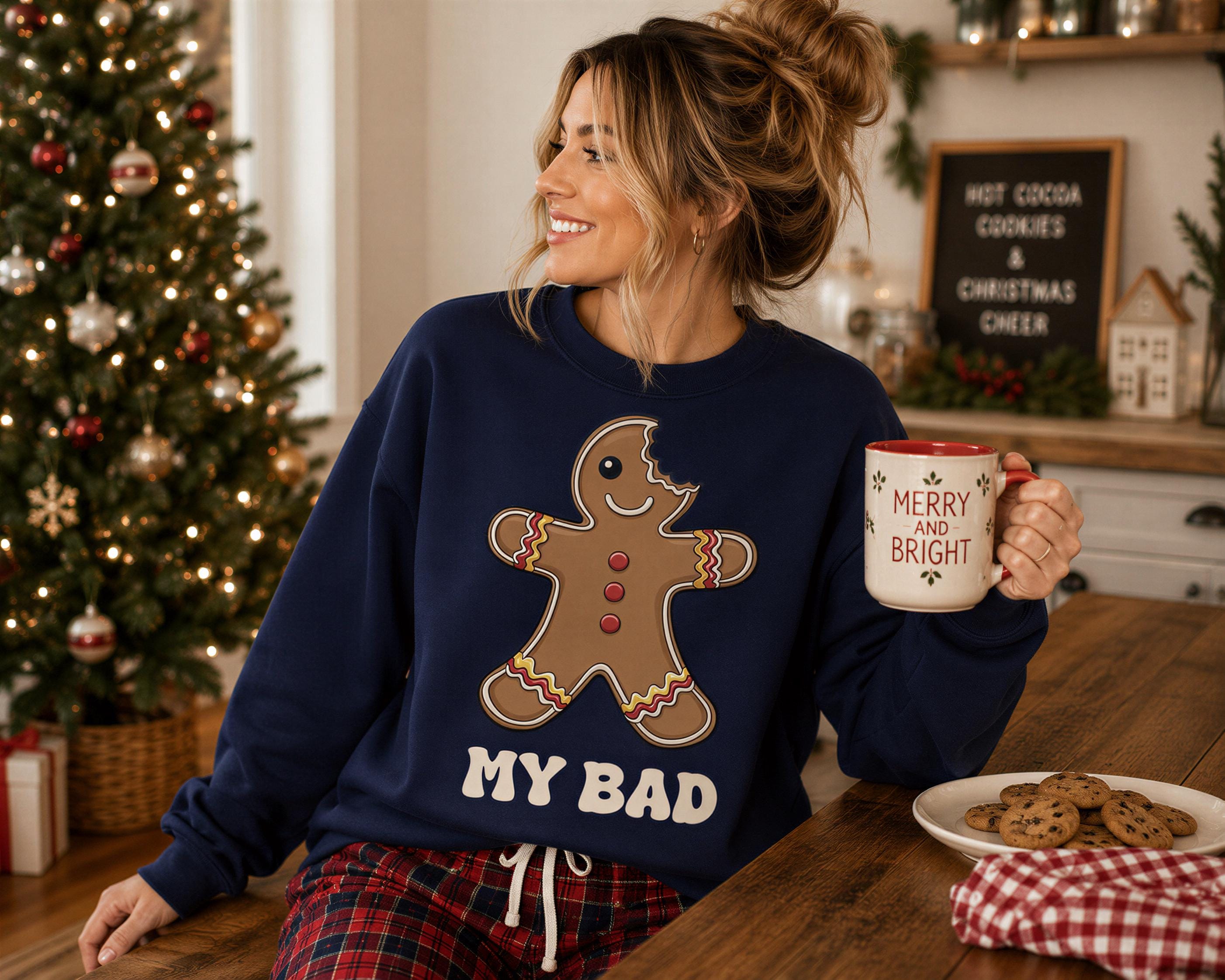 A woman wearing a blue sweatshirt with a gingerbread man graphic and the text "MY BAD" is sitting at a table with a plate of cookies and a mug.