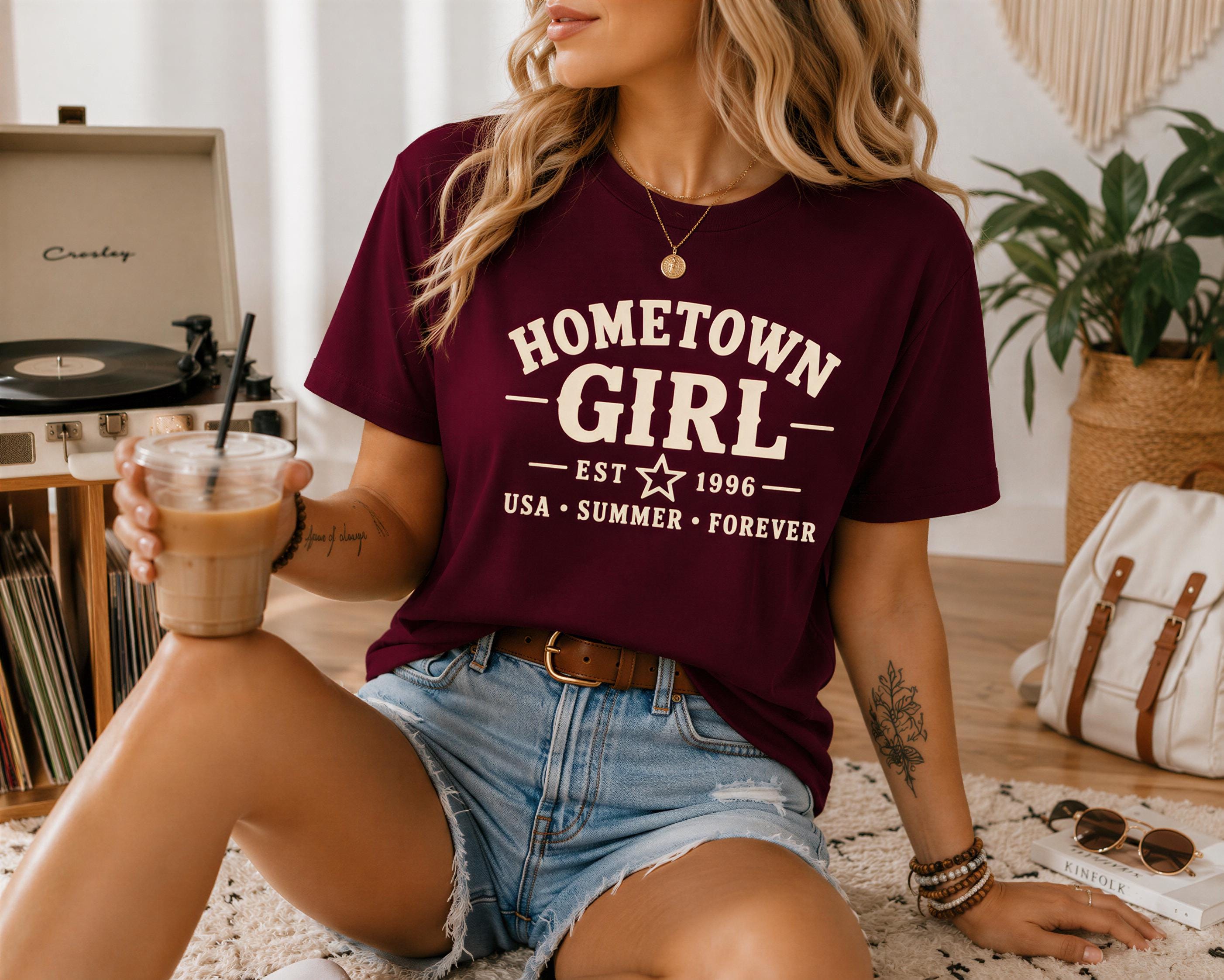 A woman wearing a burgundy t-shirt with the text "HOMETOWN GIRL" is sitting on the floor, holding a drink in her hand.