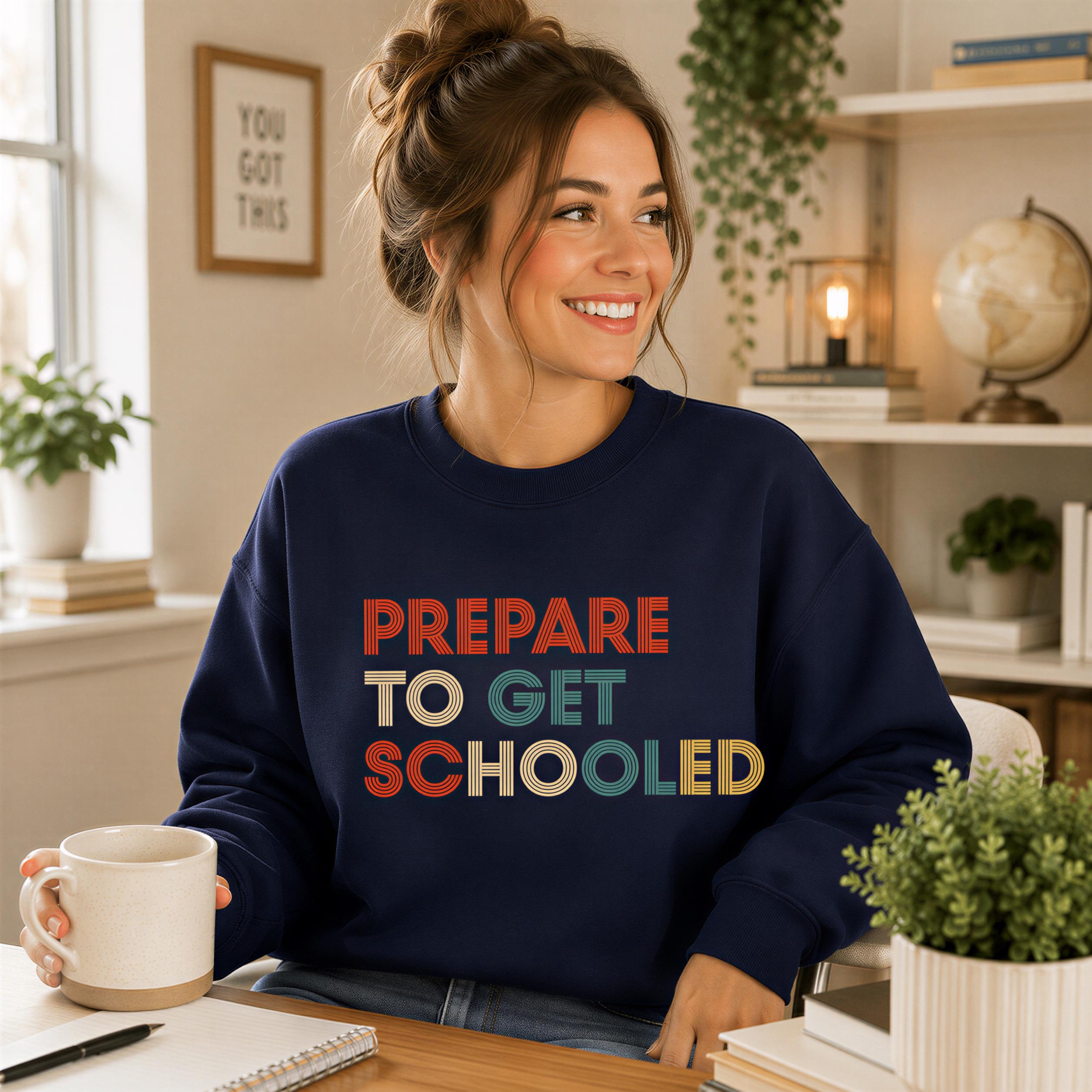 A woman wearing a navy blue sweatshirt with the text "PREPARED TO GET SCHOOLED" is sitting at a desk, smiling and holding a coffee mug.