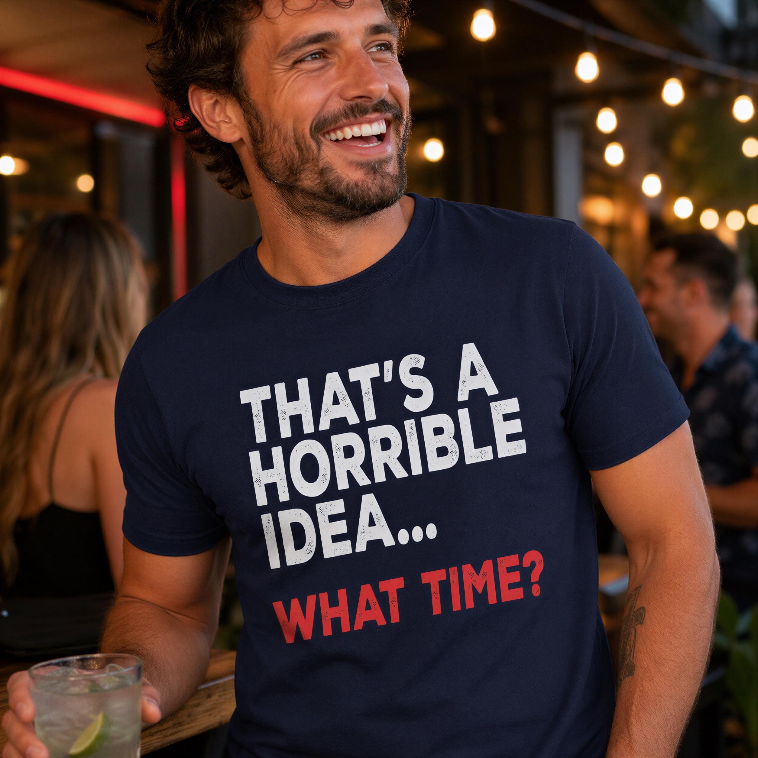 A man wearing a navy blue t-shirt with the text "THAT'S A HORRIBLE IDEA... WHAT TIME?" is smiling and holding a drink in a bar setting with other people in the background.