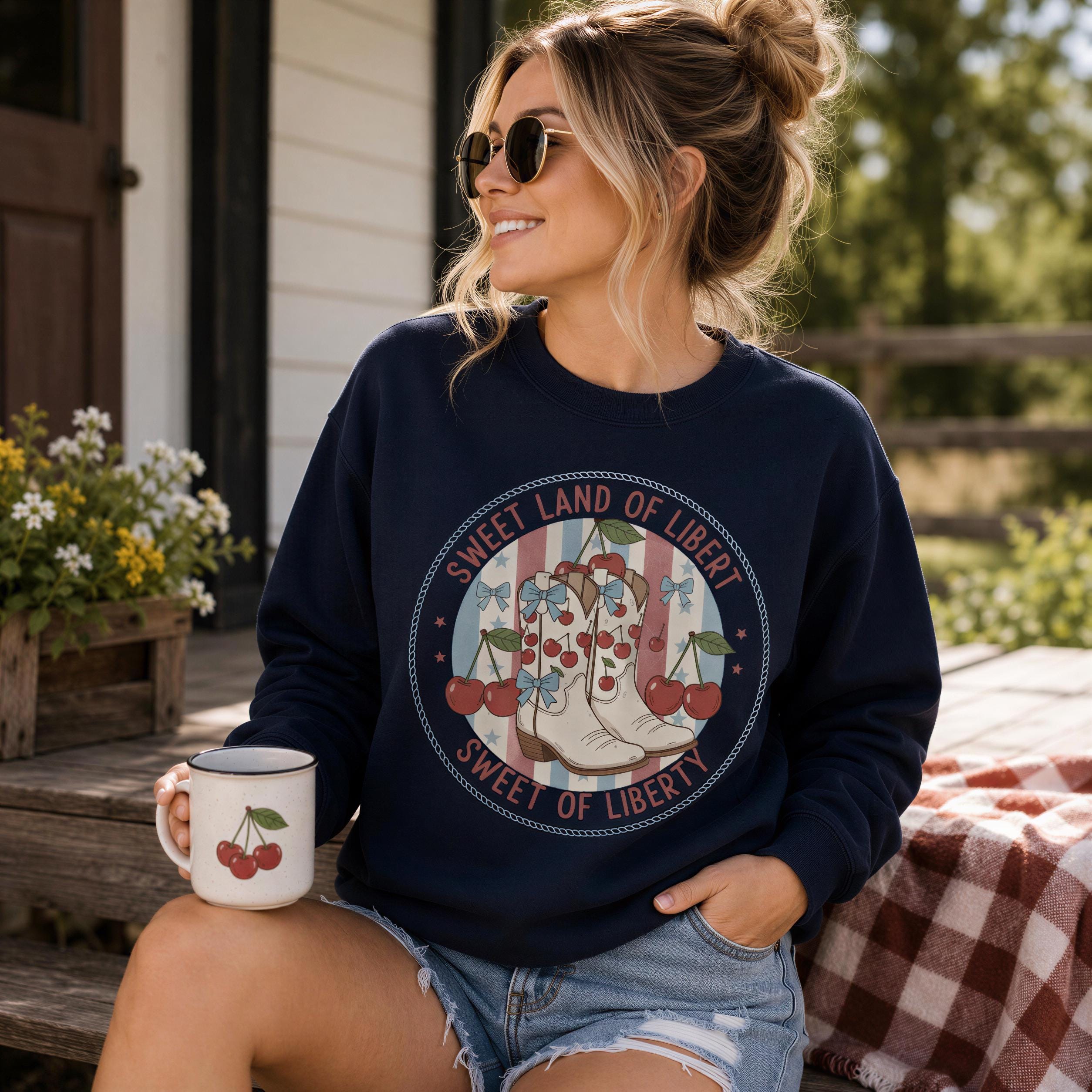 A woman wearing a navy blue sweatshirt with a graphic design of a pair of boots and cherries, sitting on a wooden bench and holding a white mug with cherries on it.