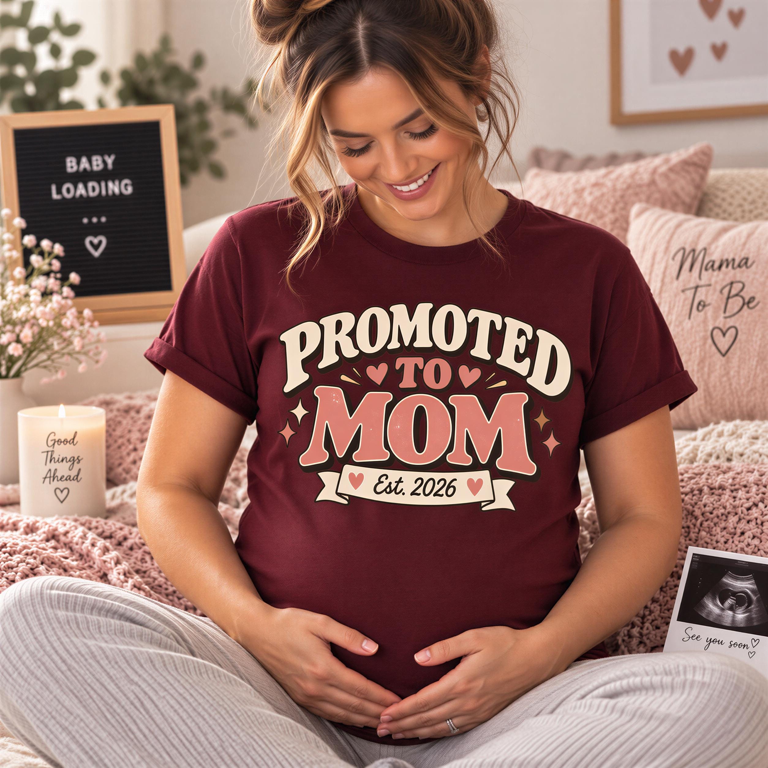 A pregnant woman wearing a maroon t-shirt with the text "Promoted to Mom" is sitting on a bed, smiling and looking down at her belly.