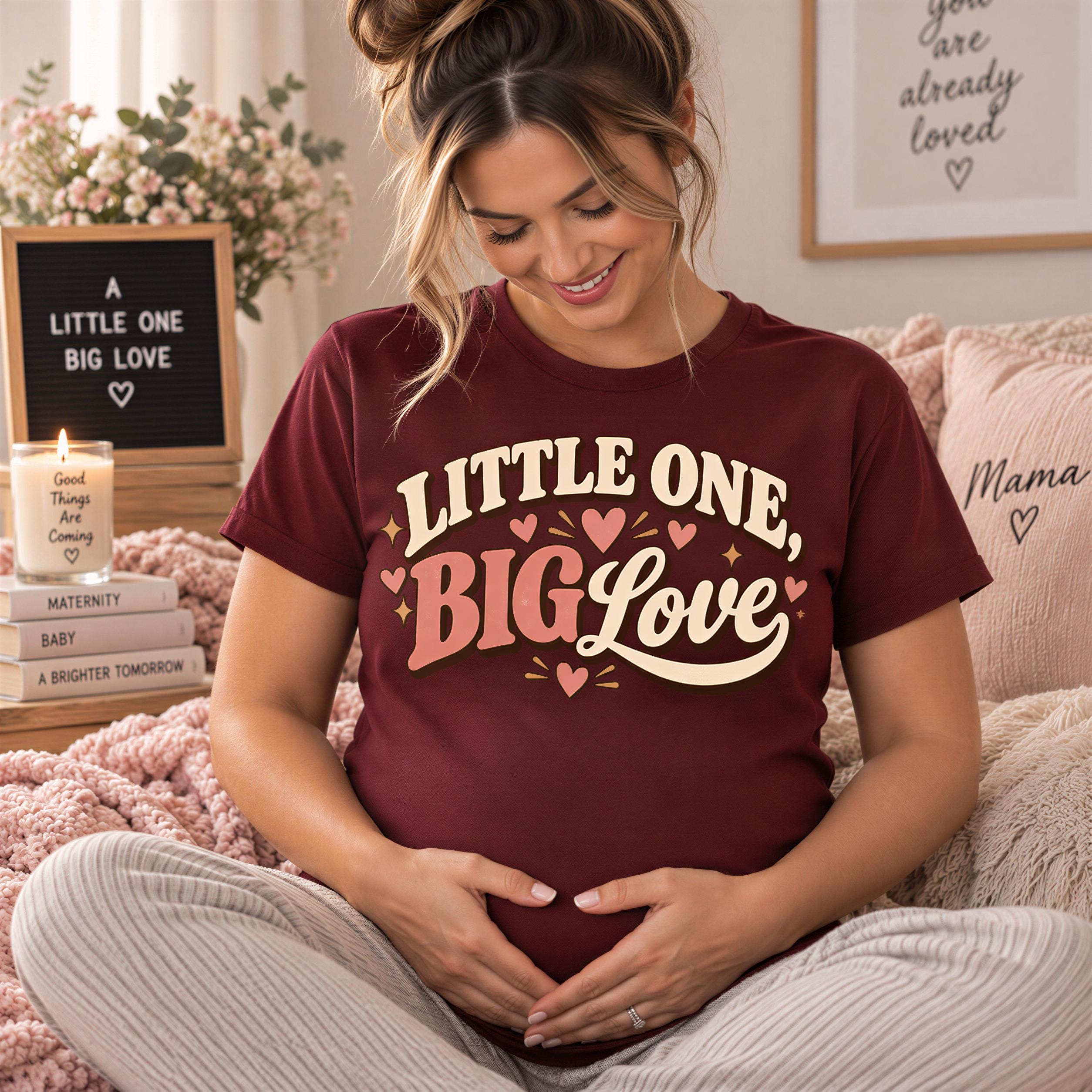 A pregnant woman wearing a burgundy t-shirt with the text "Little One Big Love" is sitting on a bed, smiling and looking down at her belly.