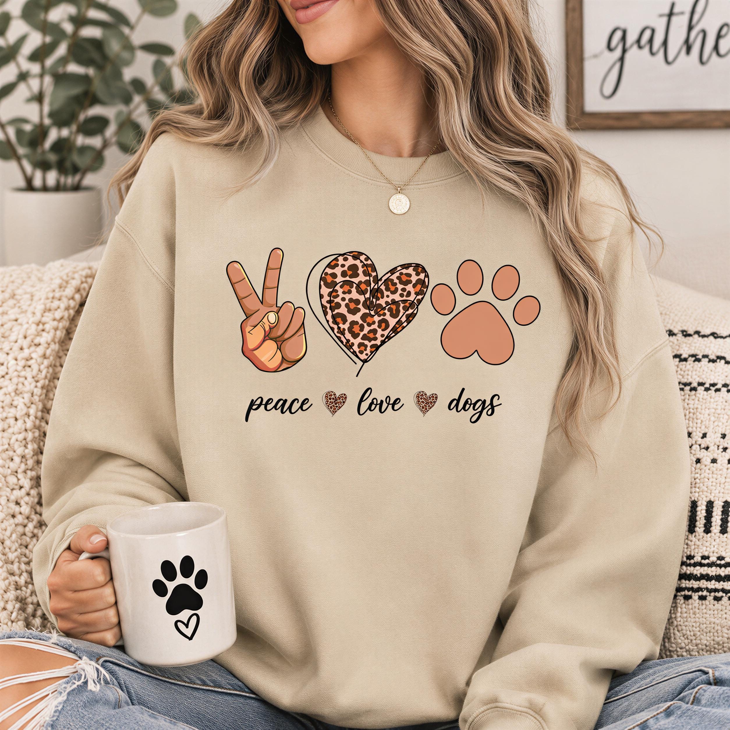 A woman wearing a beige sweatshirt with a graphic design of a peace sign, a leopard print heart, and a paw print, holding a white mug with a paw print design.