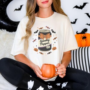 Pode incluir: Camiseta creme com um gráfico temático de Halloween. O design inclui uma lata com a etiqueta "Spooky Pepper" com um laço, rodeada por morcegos, abóboras e doces. Uma caneca laranja em forma de abóbora está sendo segurada.