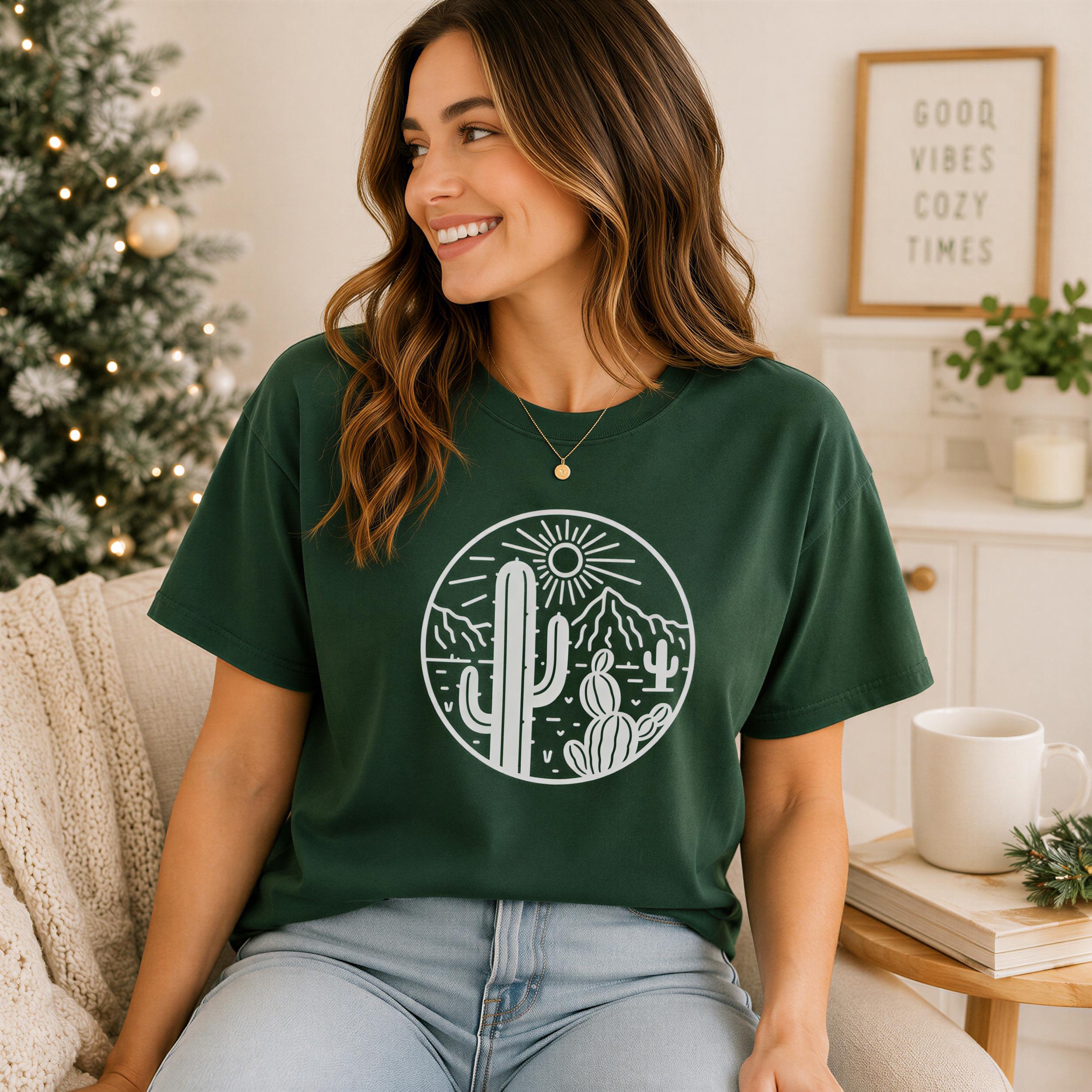 A woman wearing a green t-shirt with a desert-themed graphic is sitting on a couch, smiling and looking to the side.
