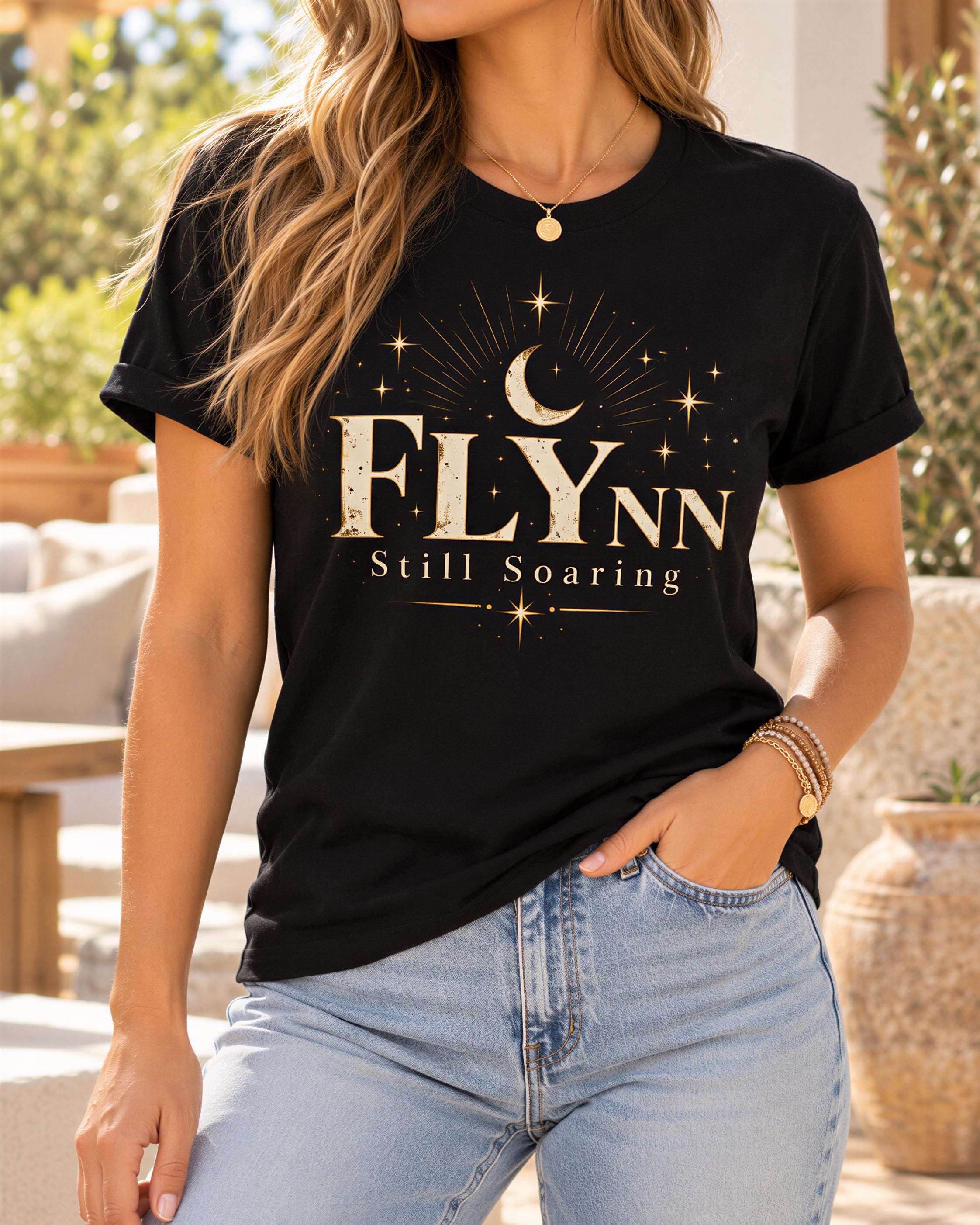 A woman wearing a black t-shirt with a graphic design and text, standing in an outdoor setting with plants in the background.