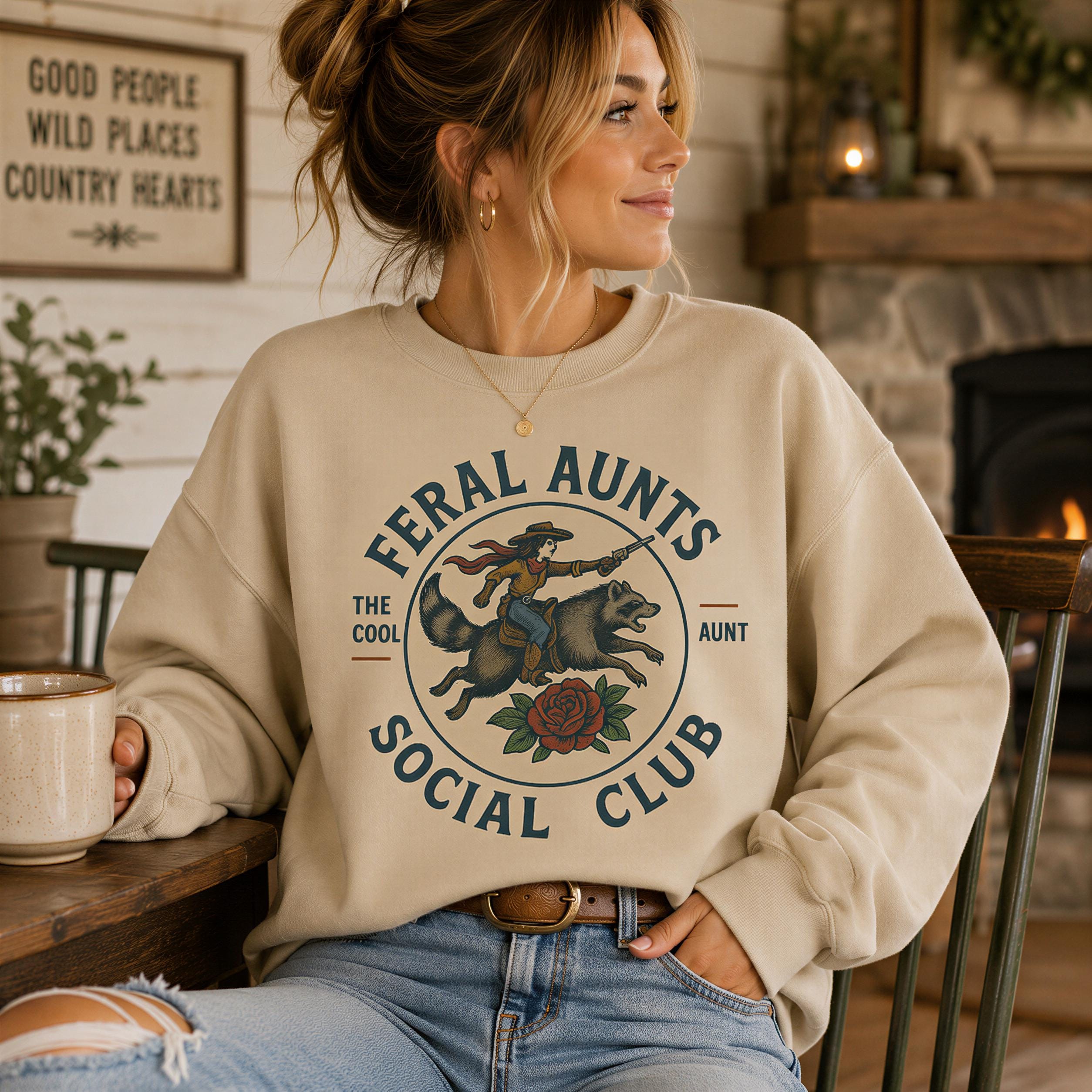 A woman wearing a beige sweatshirt with a graphic design and text, sitting in a room with a fireplace and a sign that says "Good people wild places country hearts".
