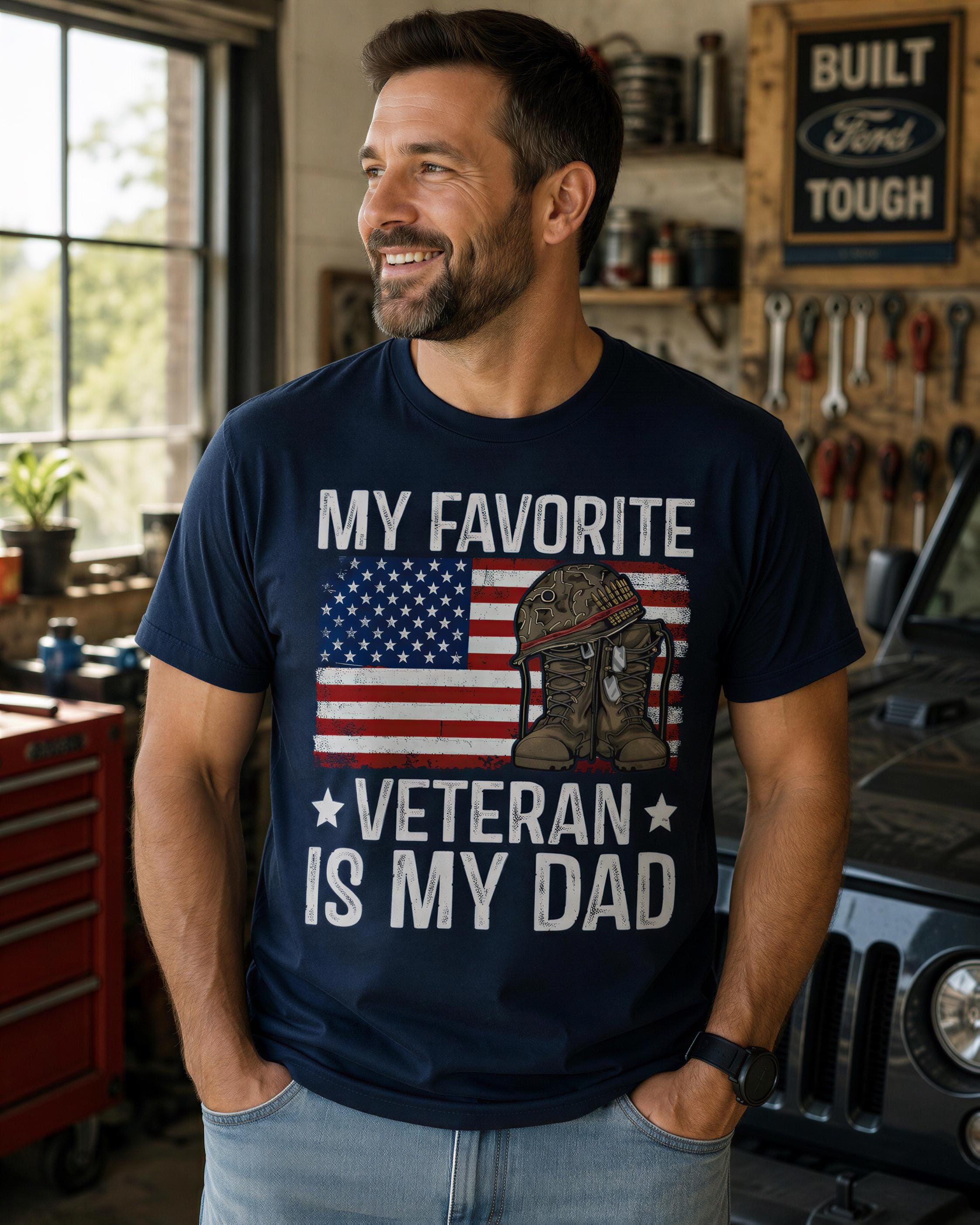 A man wearing a navy blue t-shirt with the text "My favorite Veteran is my dad" and an American flag graphic, smiling and looking to the side.