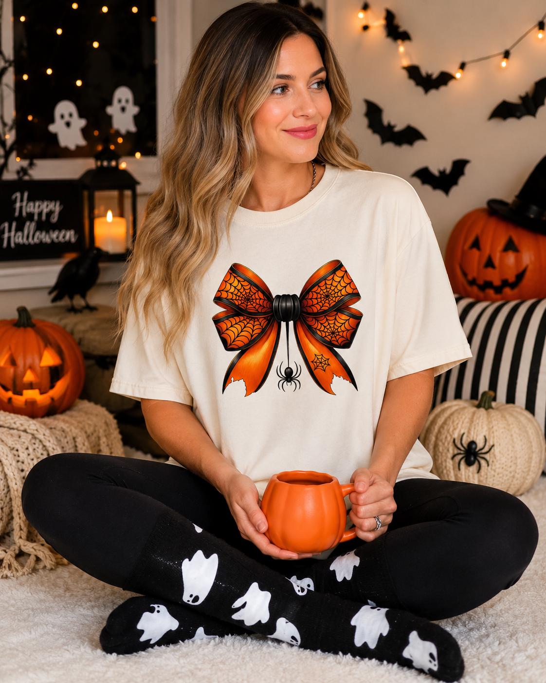 A woman sits on the floor, wearing a white t-shirt with a large orange bow and black pants with ghost and spider designs. She holds a mug in her hands. Halloween decorations, including jack-o-lanterns and bats, are visible in the background.