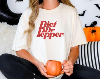 Puede incluir: Camiseta color crema con las palabras "Diet Dr Pepper" en rojo. La persona sostiene una taza naranja con forma de calabaza. El fondo incluye decoraciones de Halloween como murciélagos y una calabaza.