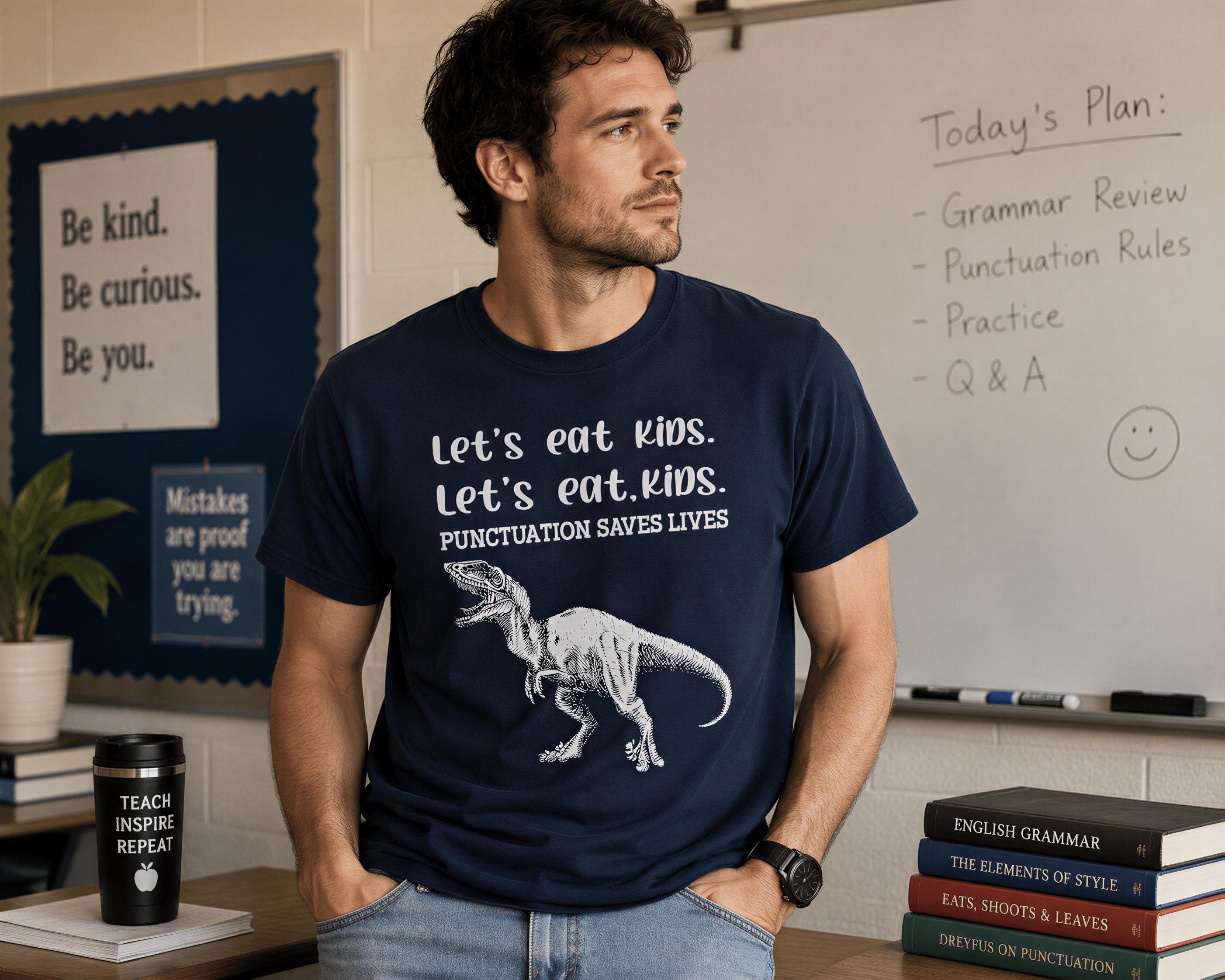 A man wearing a blue t-shirt with a dinosaur graphic and text stands in a classroom setting, with a whiteboard and bookshelves visible in the background.