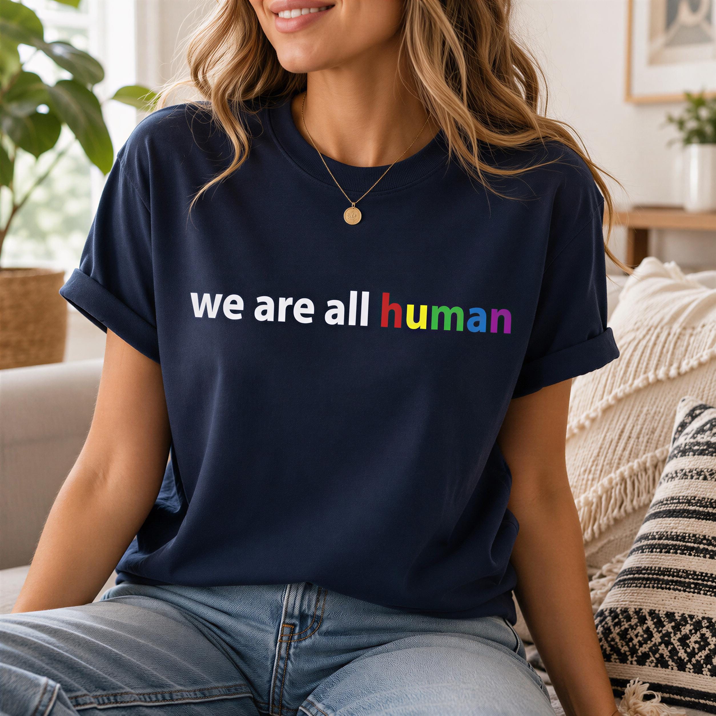 A woman wearing a navy blue t-shirt with the text "we are all human" printed on it, sitting on a couch.