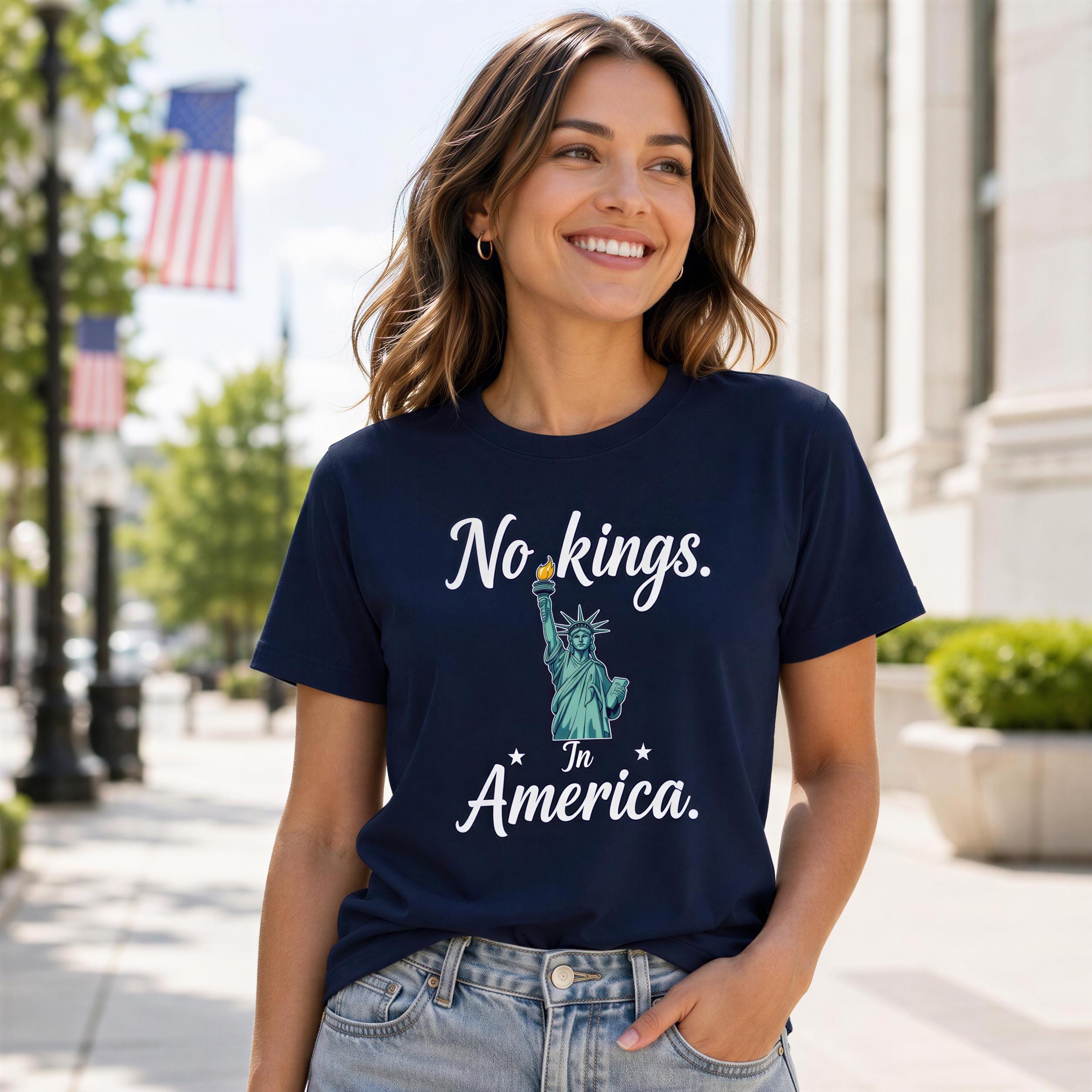 A woman wearing a navy blue t-shirt with the text "No kings in America" printed on it, smiling and standing on a city street.