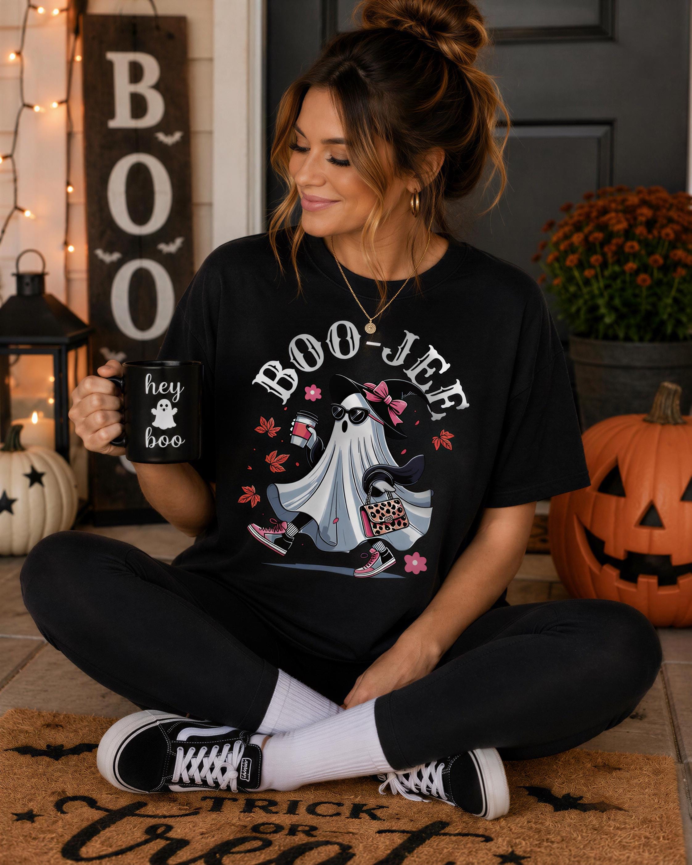 A woman sitting on the floor, wearing a black t-shirt with a graphic of a ghost and holding a mug with the text "hey ghost" printed on it. She is surrounded by Halloween decorations, including a pumpkin and a "BOO JEF" sign.