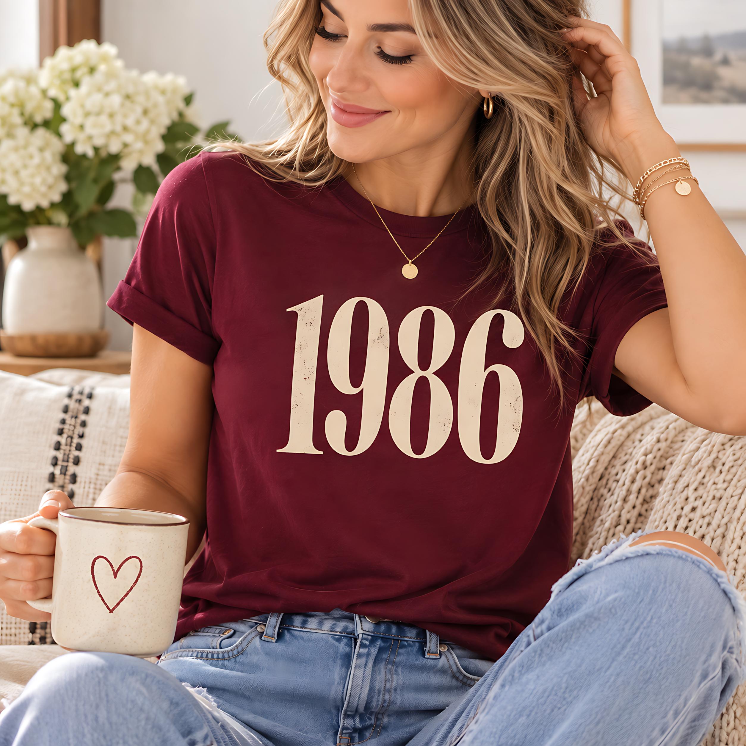 A woman wearing a burgundy t-shirt with the number 1986 and holding a coffee mug is sitting on a couch.