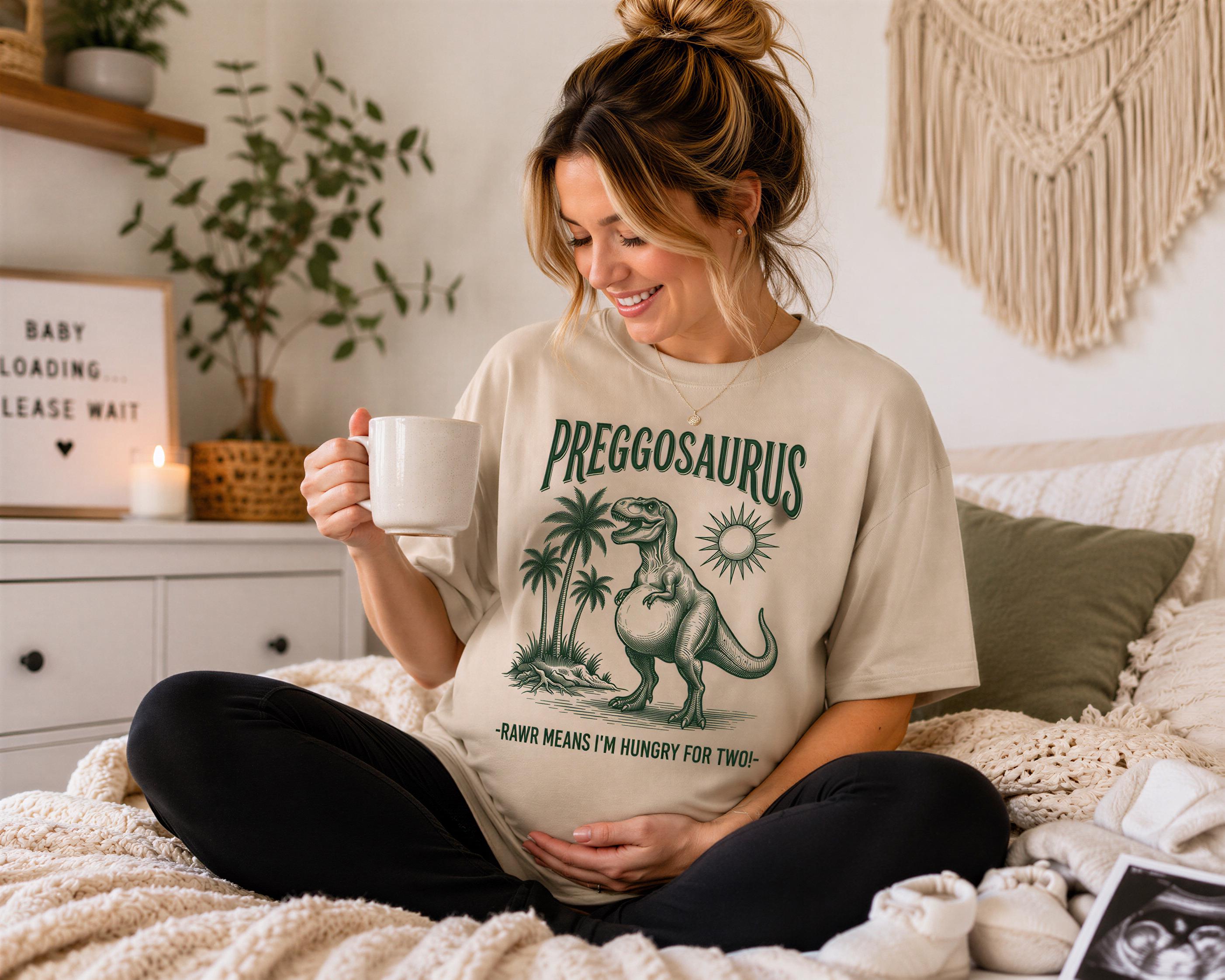 A pregnant woman sitting on a bed, holding a mug and smiling, wearing a t-shirt with a dinosaur graphic and text.