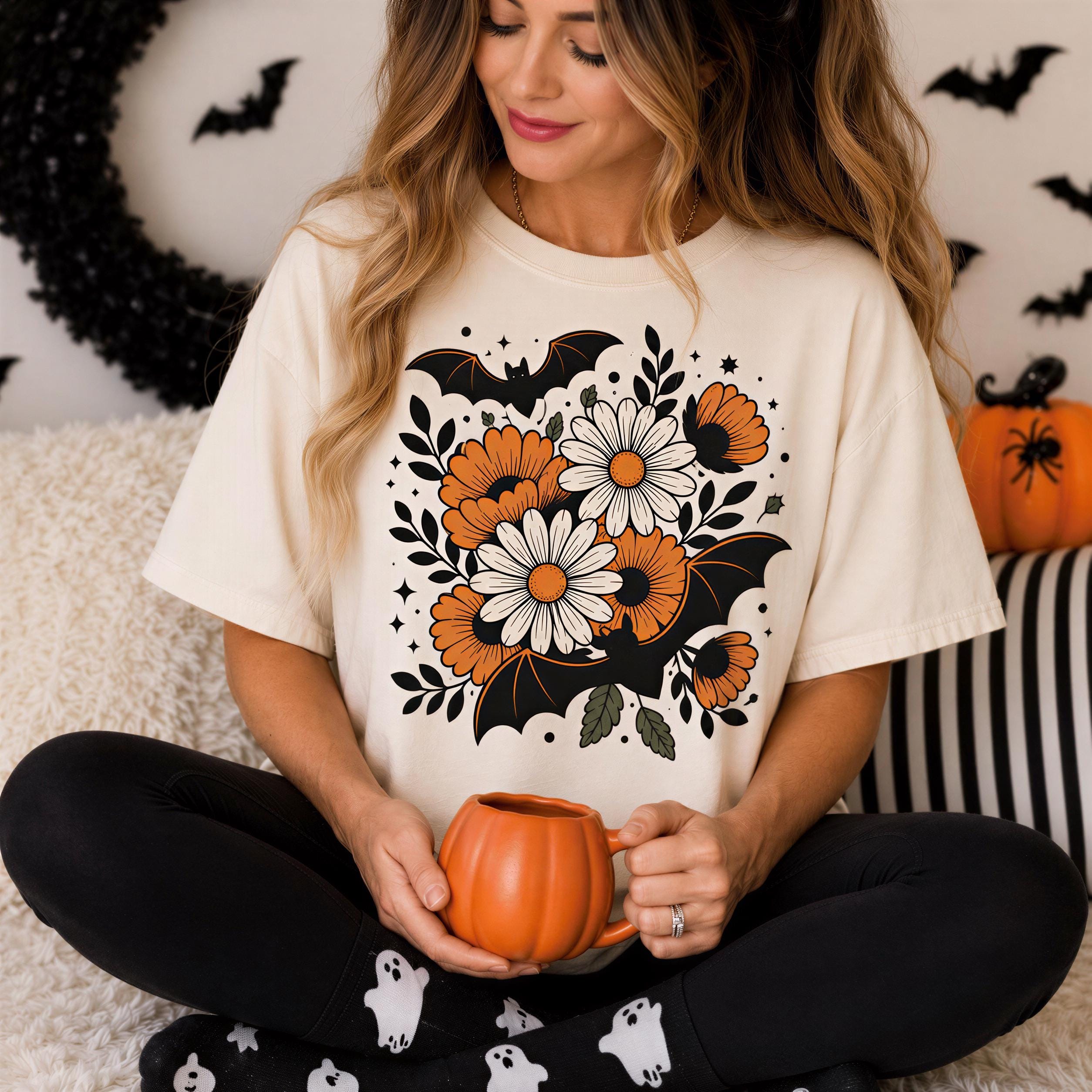 A woman is sitting on the floor, holding a pumpkin-shaped mug, with a floral t-shirt featuring a bat design and a pumpkin decoration.