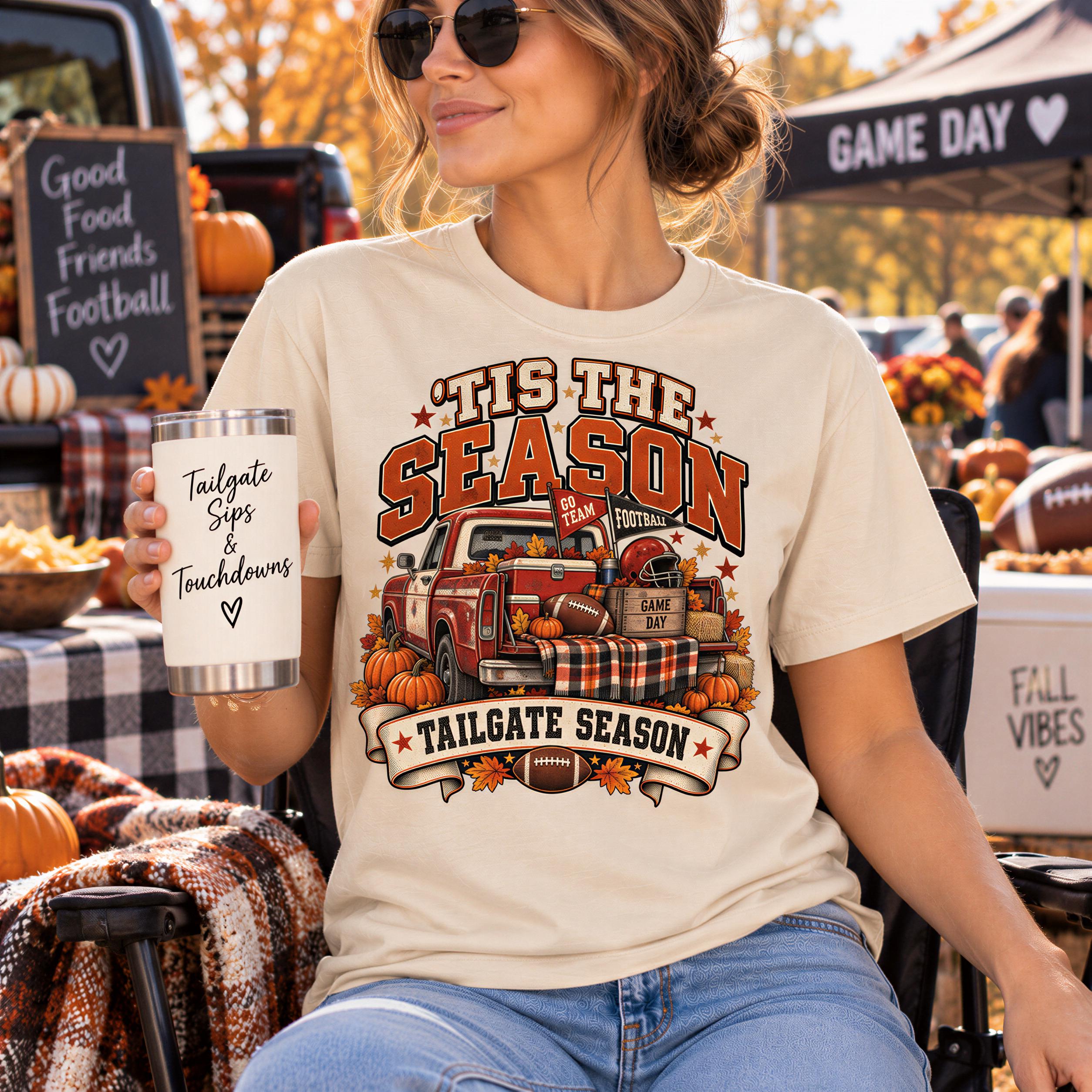 A woman wearing a t-shirt with a graphic design of a red truck and autumn decorations, holding a cup with the text "Tis the season Football" and "Tailgate Sips & Touchdowns" printed on it. She is sitting in a chair at an outdoor event with a sign that says "Good Food Friends Football" and other fall decorations around her.