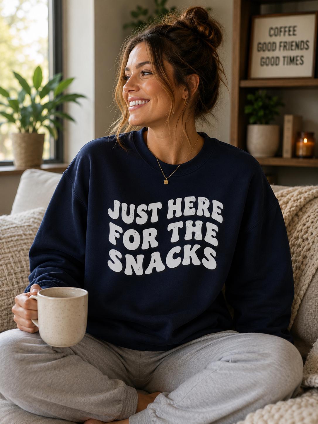 A woman sitting on a couch, wearing a navy blue sweatshirt with the text "Just here for the snacks" printed on it. She is holding a coffee mug and smiling.