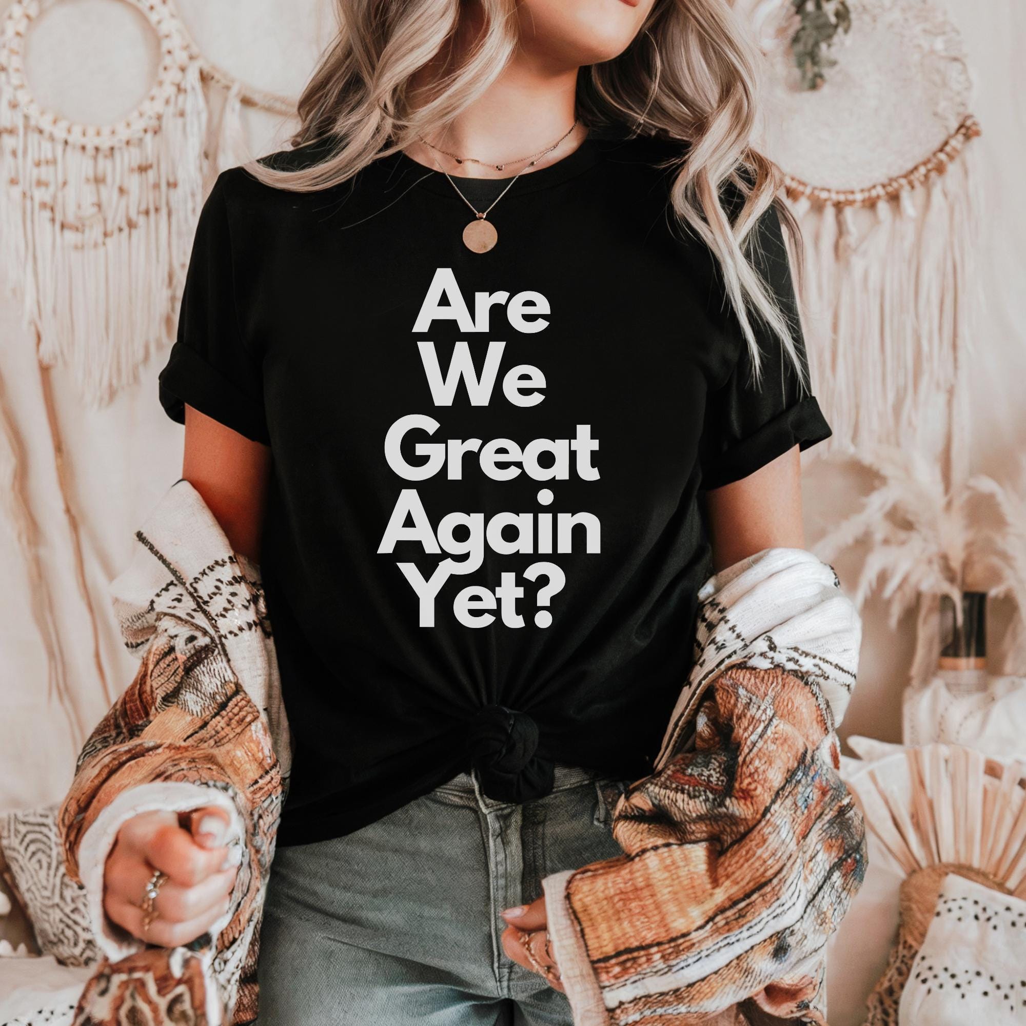 A woman wearing a black t-shirt with the text "Are We Great Again Yet?" stands in front of a beige wall with a dreamcatcher and a woven basket.