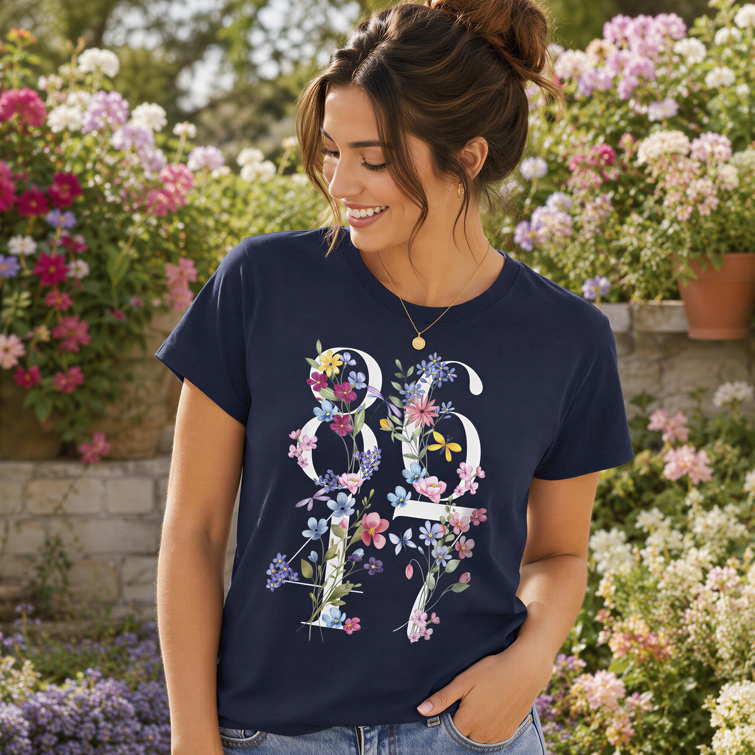 A woman wearing a navy blue t-shirt with a floral design stands in a garden surrounded by colorful flowers.
