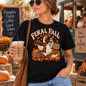 May include: A black t-shirt featuring a graphic of a goose wearing a plaid scarf, holding a coffee cup, and standing near pumpkins and a stack of books. The text reads FERAL FALL CLUB and SILLY GOOSE. The design includes autumn leaves.