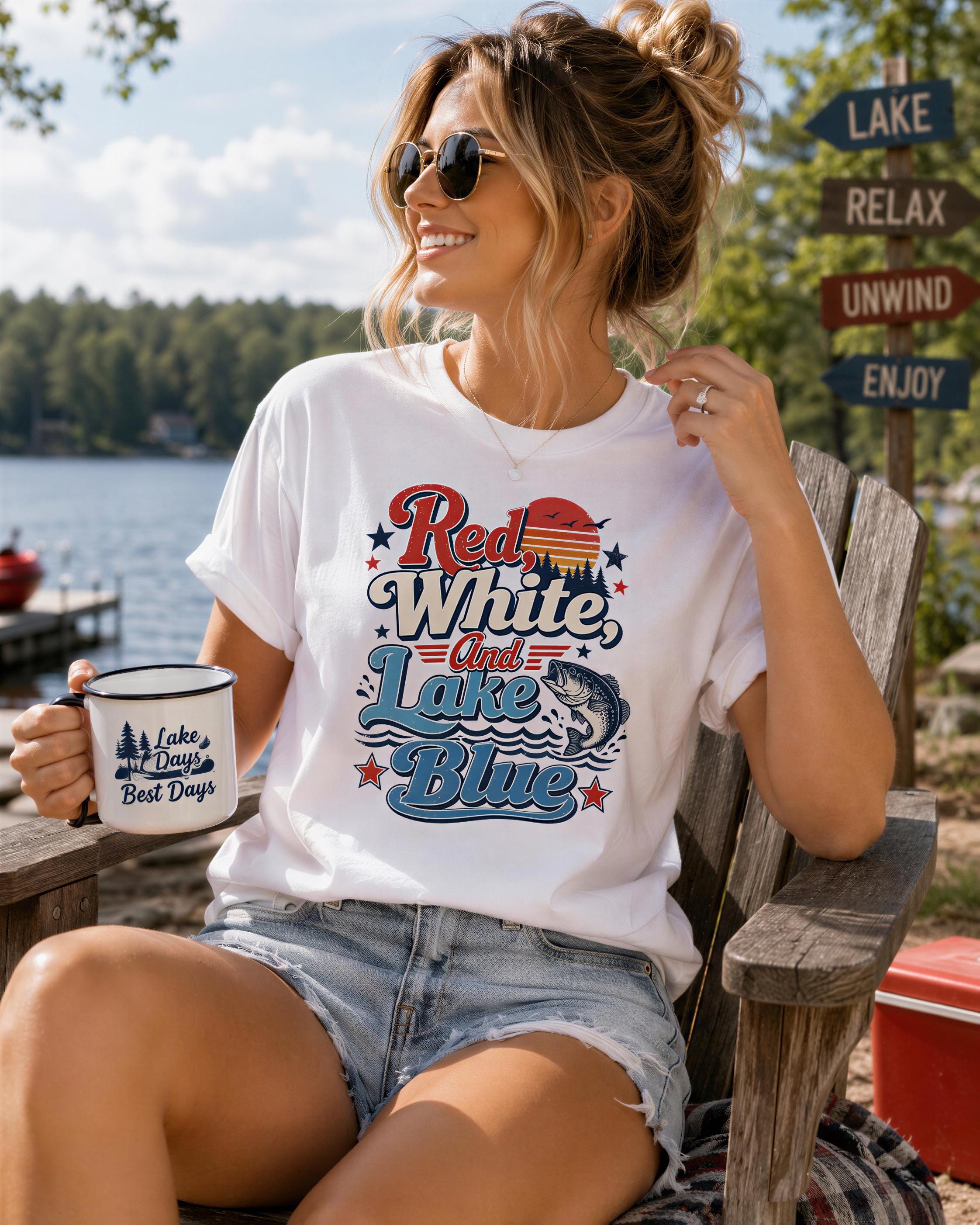 A woman wearing a white t-shirt with a graphic design and sunglasses is sitting on a wooden chair by a lake, holding a mug.
