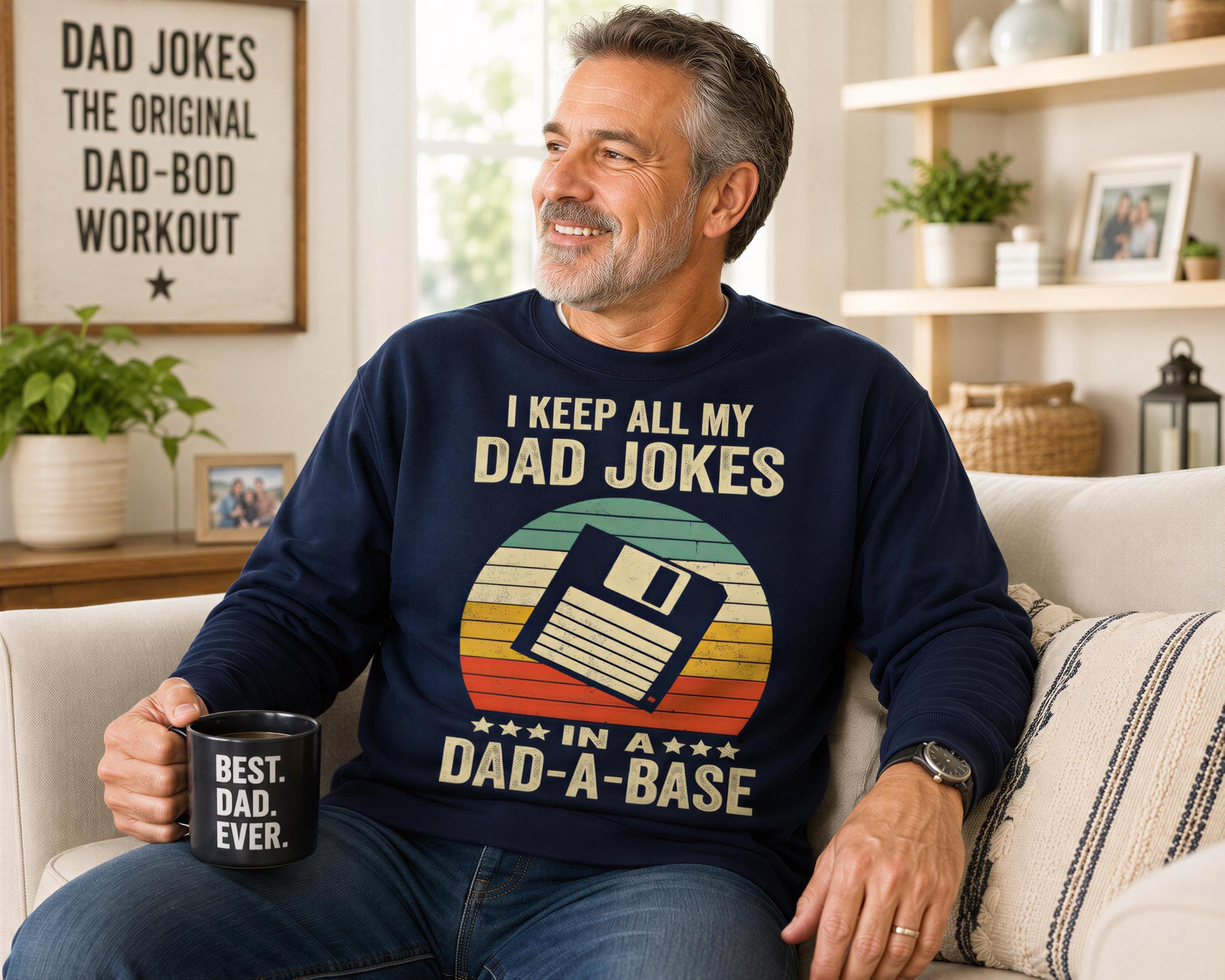 A man sitting on a couch, wearing a blue sweatshirt with a humorous message about keeping all his dad jokes in a "dad-a-base" style. He is holding a coffee mug with the same message printed on it.