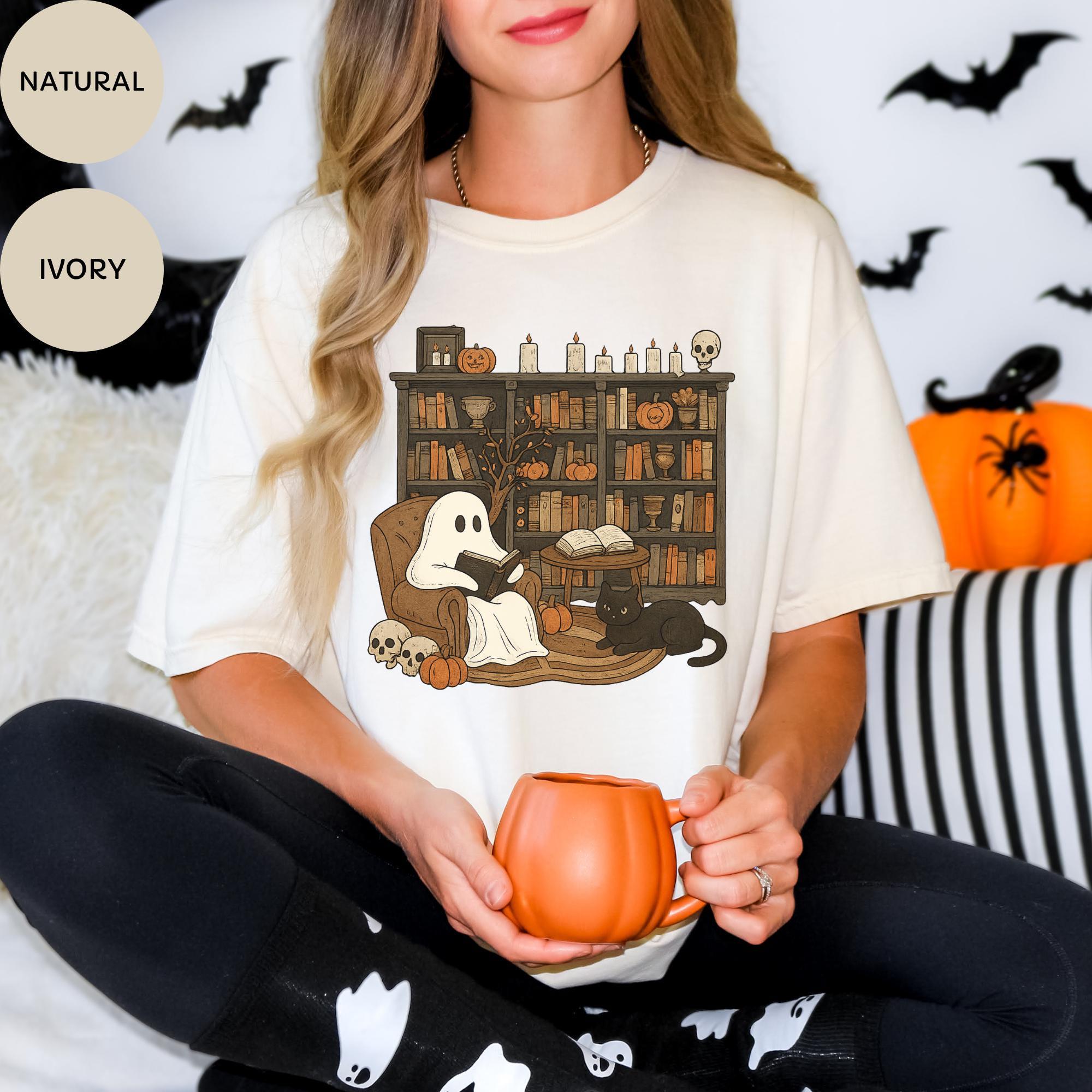 A woman wearing a white t-shirt with a graphic of a ghost sitting in a chair surrounded by books and pumpkins, holding a pumpkin-shaped mug.