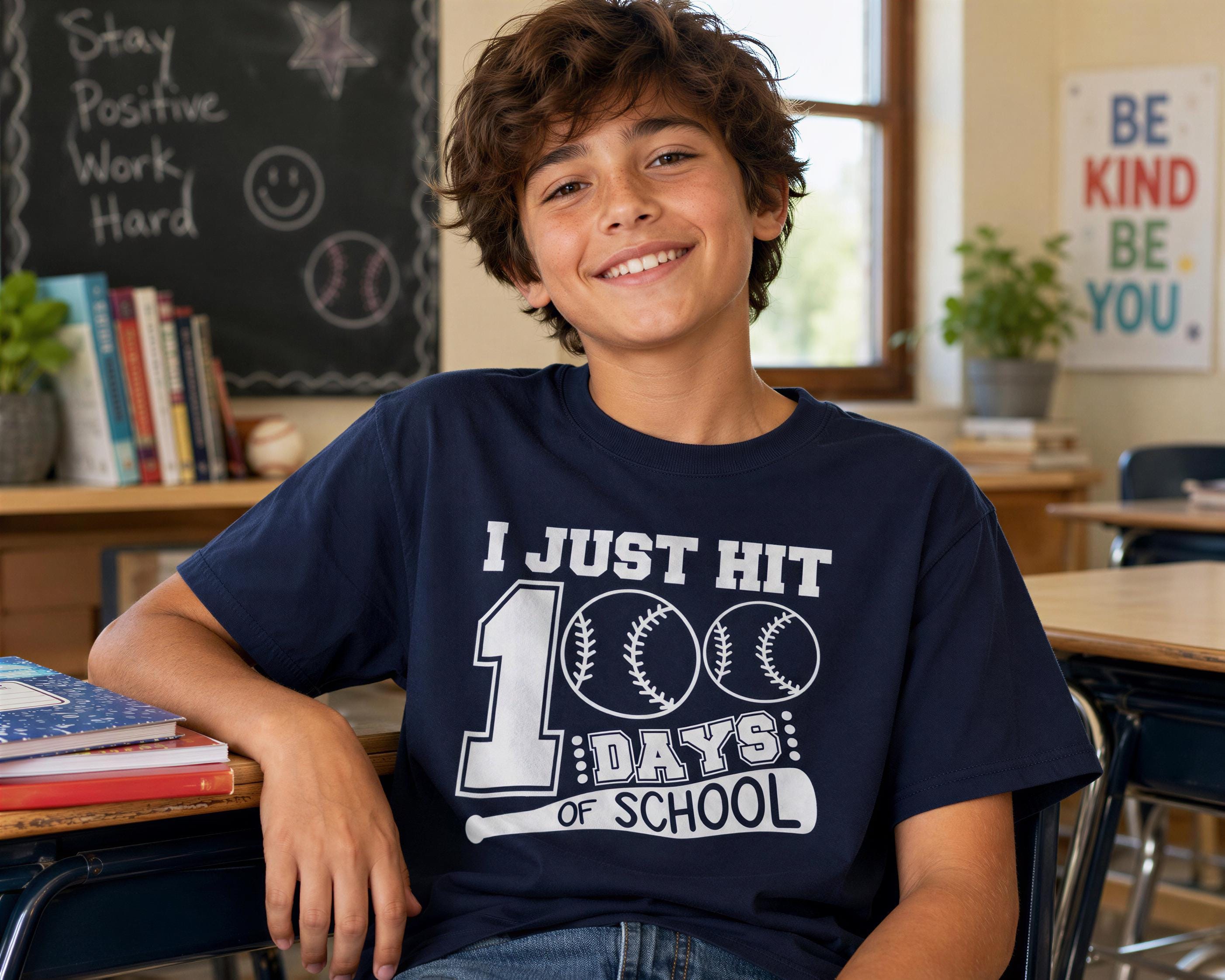 A smiling young boy wearing a blue t-shirt with a baseball graphic and text, sitting in a classroom setting with a chalkboard and bookshelves in the background.
