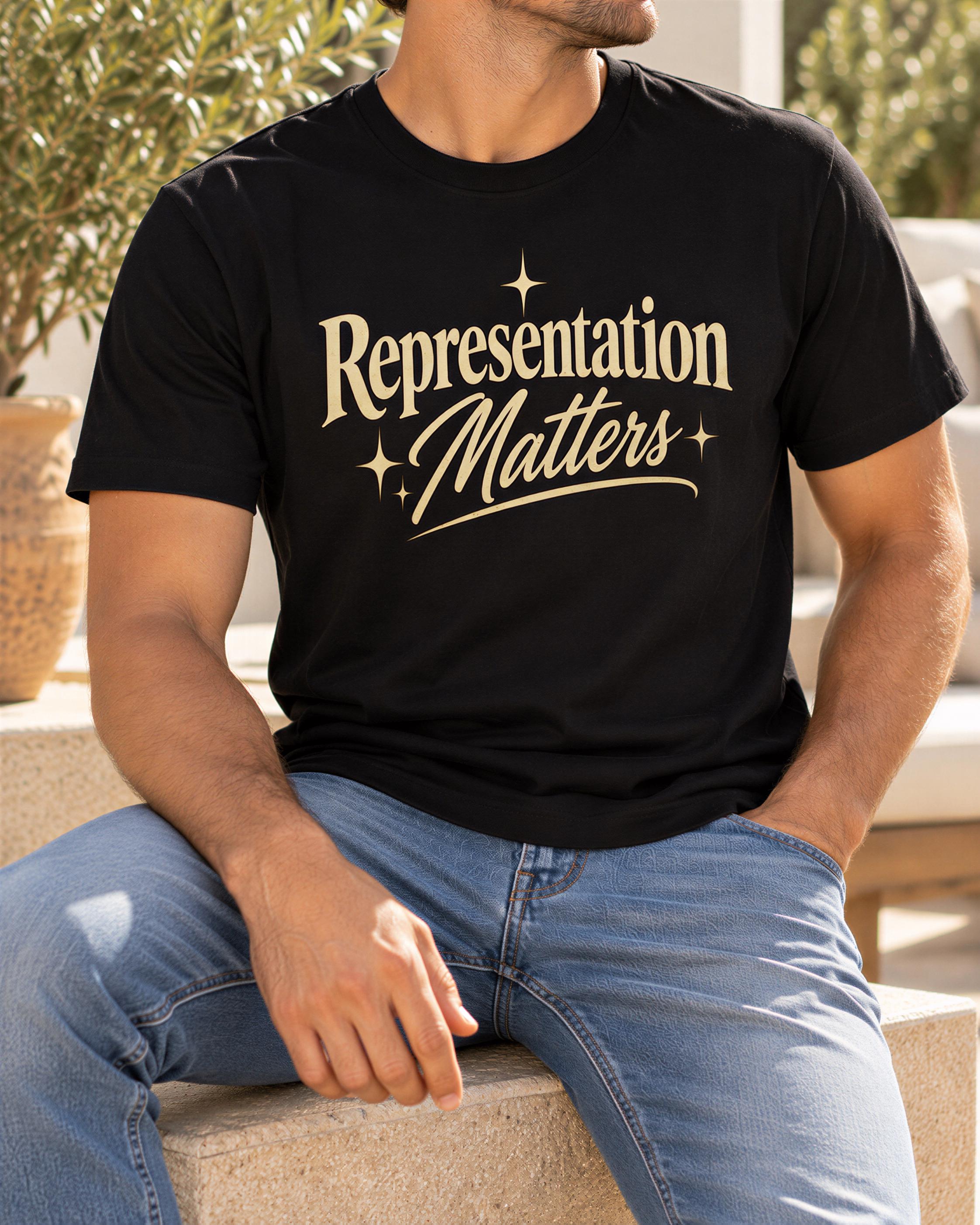 A man wearing a black t-shirt with the text "Representation Matters" printed on it, sitting on a bench outdoors.
