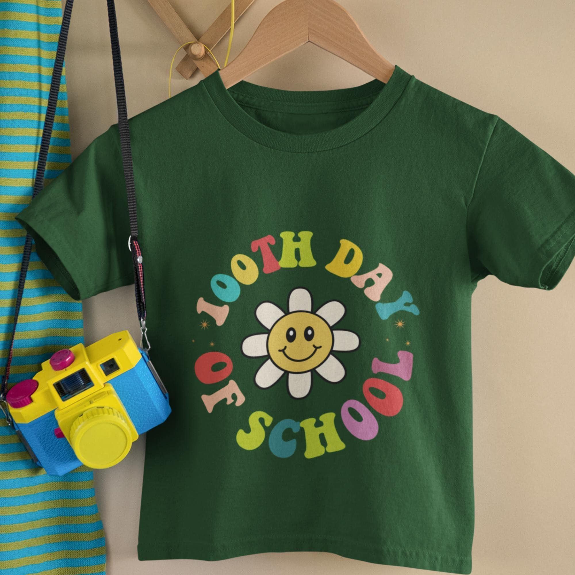 A green t-shirt with a colorful flower design and the text "I THINK DAY OF SCHOOL" printed on it, along with a yellow camera hanging on a wooden hanger.