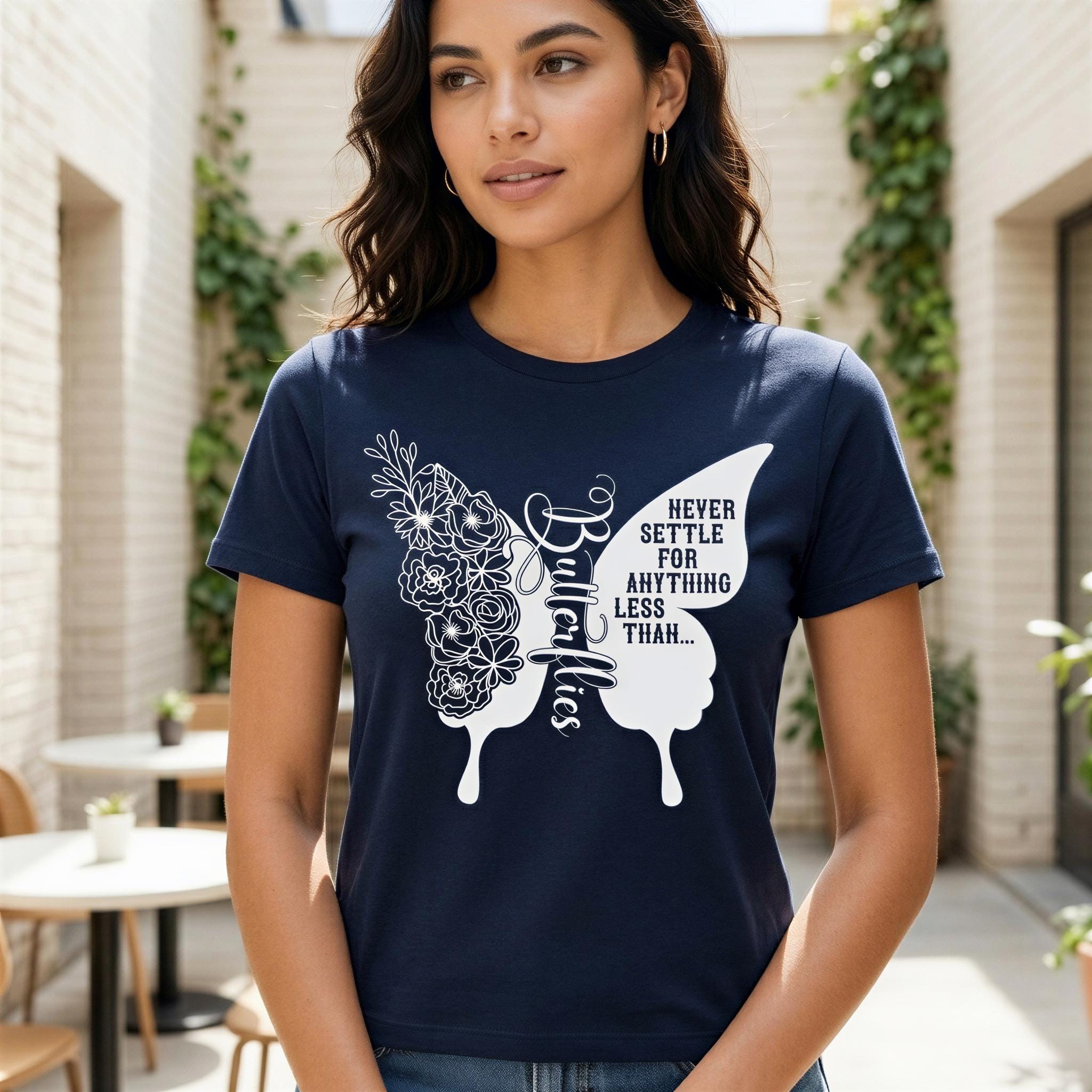 A woman wearing a navy blue t-shirt with a white butterfly design and text printed on it, standing in an indoor setting with plants and tables visible in the background.