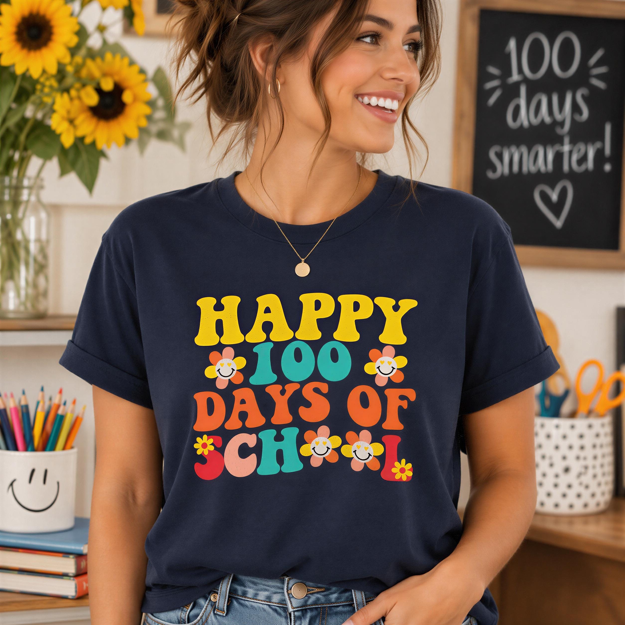 A woman wearing a blue t-shirt with a colorful graphic design that says "HAPPY 100 DAYS OF SCHOOL" and features various school-related elements such as flowers and pencils.