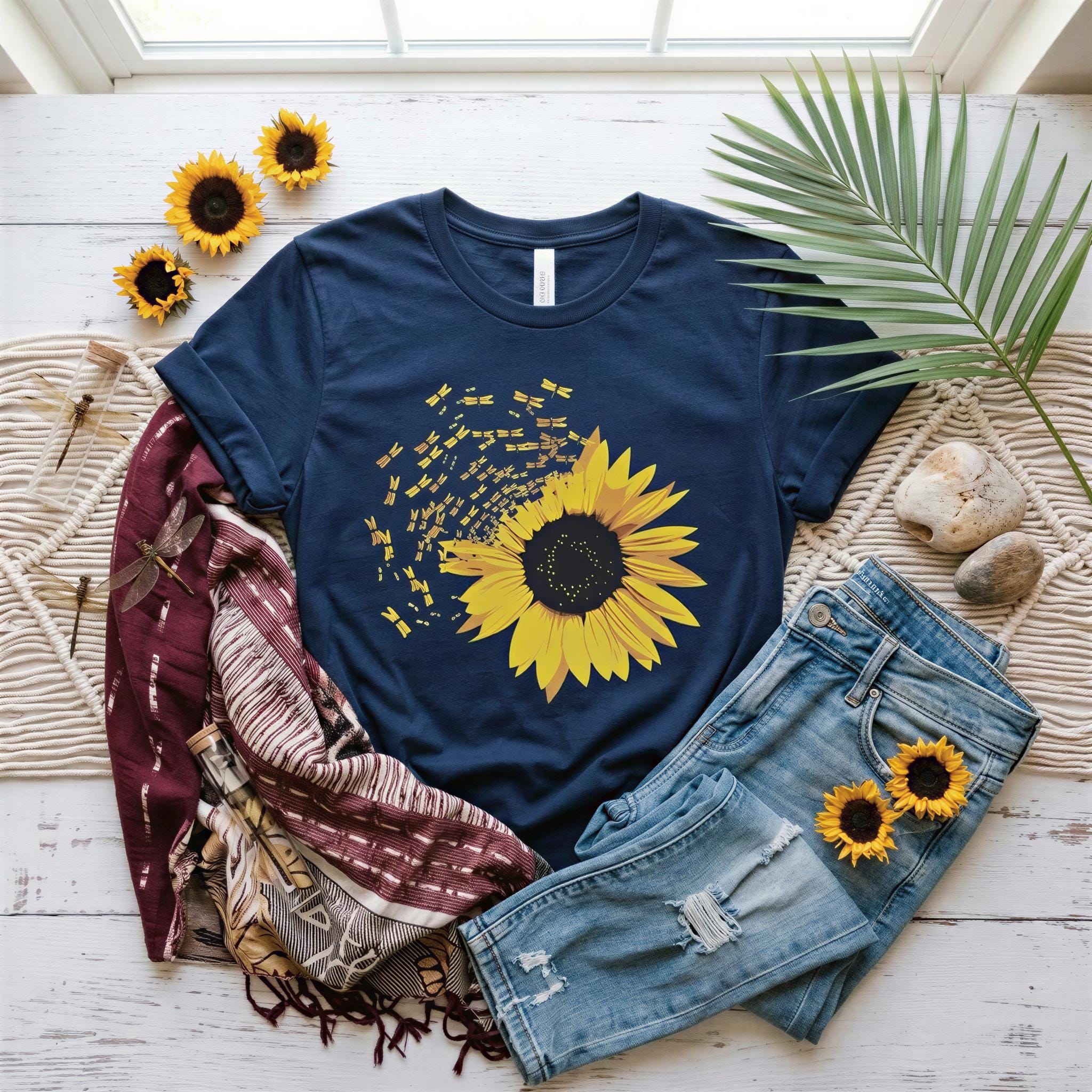 A navy blue t-shirt with a yellow sunflower design, a burgundy shawl, a pair of blue jeans, and a green plant are arranged on a white wooden surface.