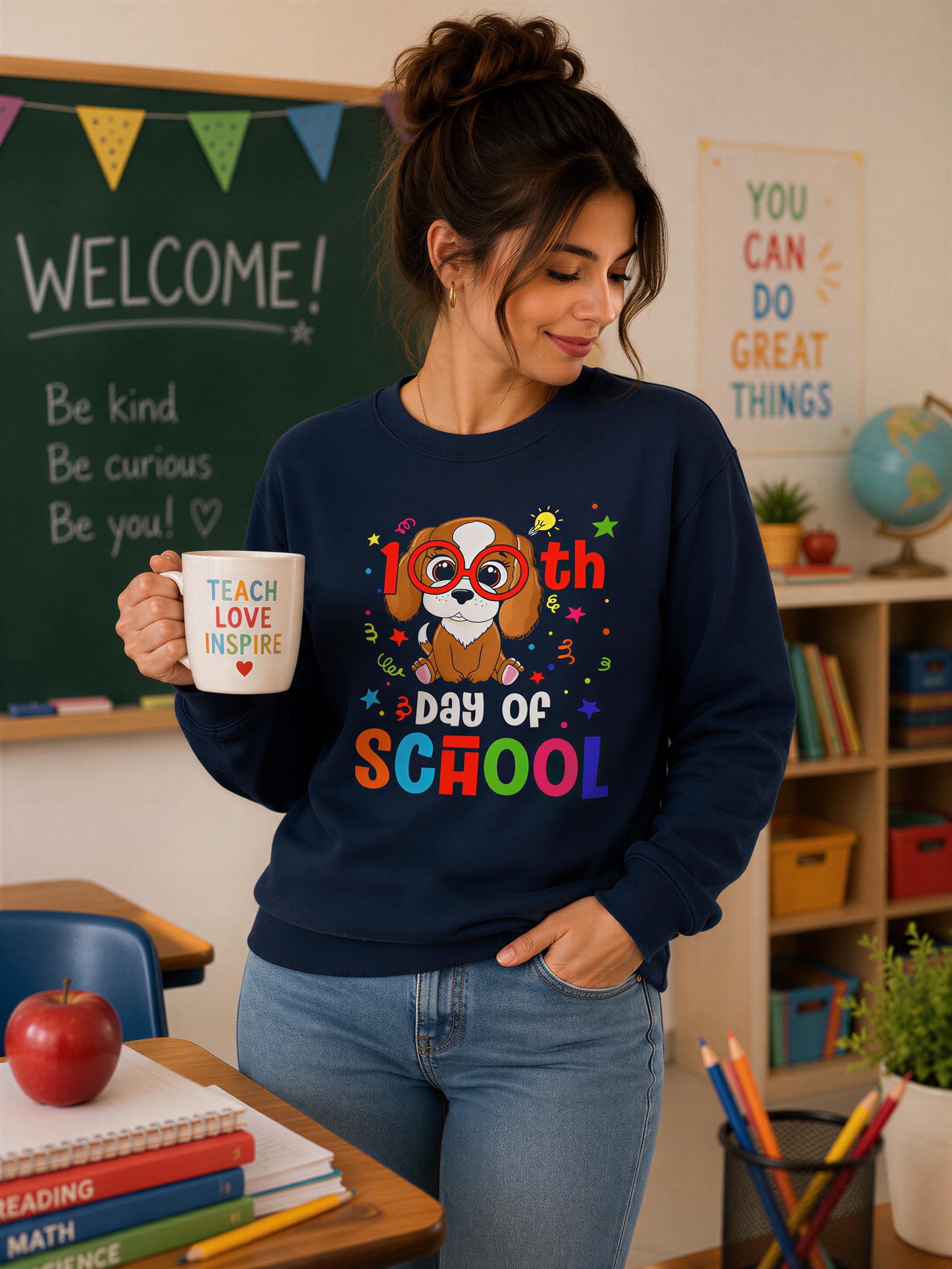 A woman wearing a blue sweatshirt with a cartoon dog and text stands in a classroom, holding a coffee mug with a design that says "Teach Love Inspire Day of School".