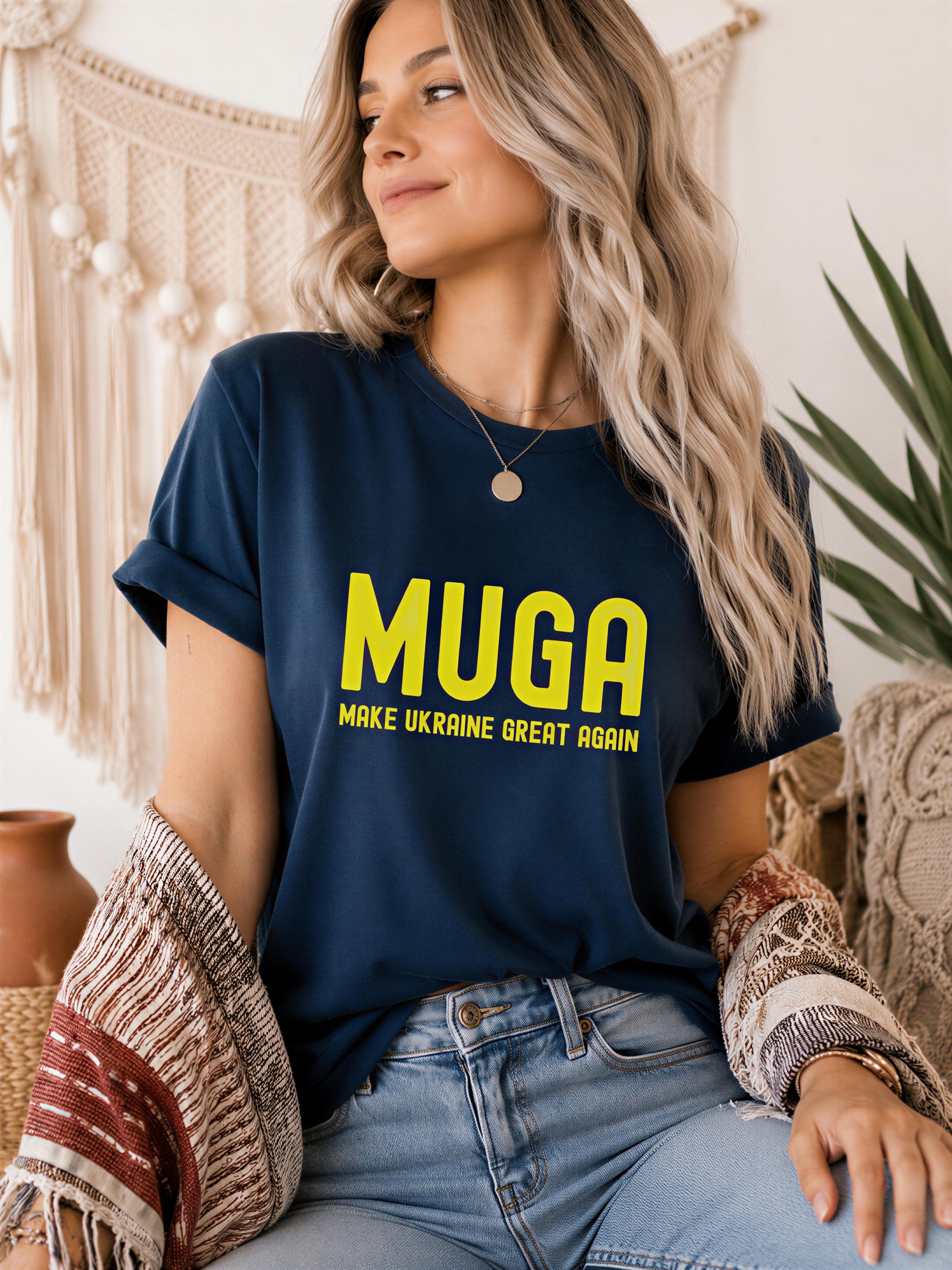 A woman with long blonde hair wearing a navy blue t-shirt with the text "MUGA" and "MAKE UKRAINE GREAT AGAIN" printed on it, sitting in a room with a plant in the background.