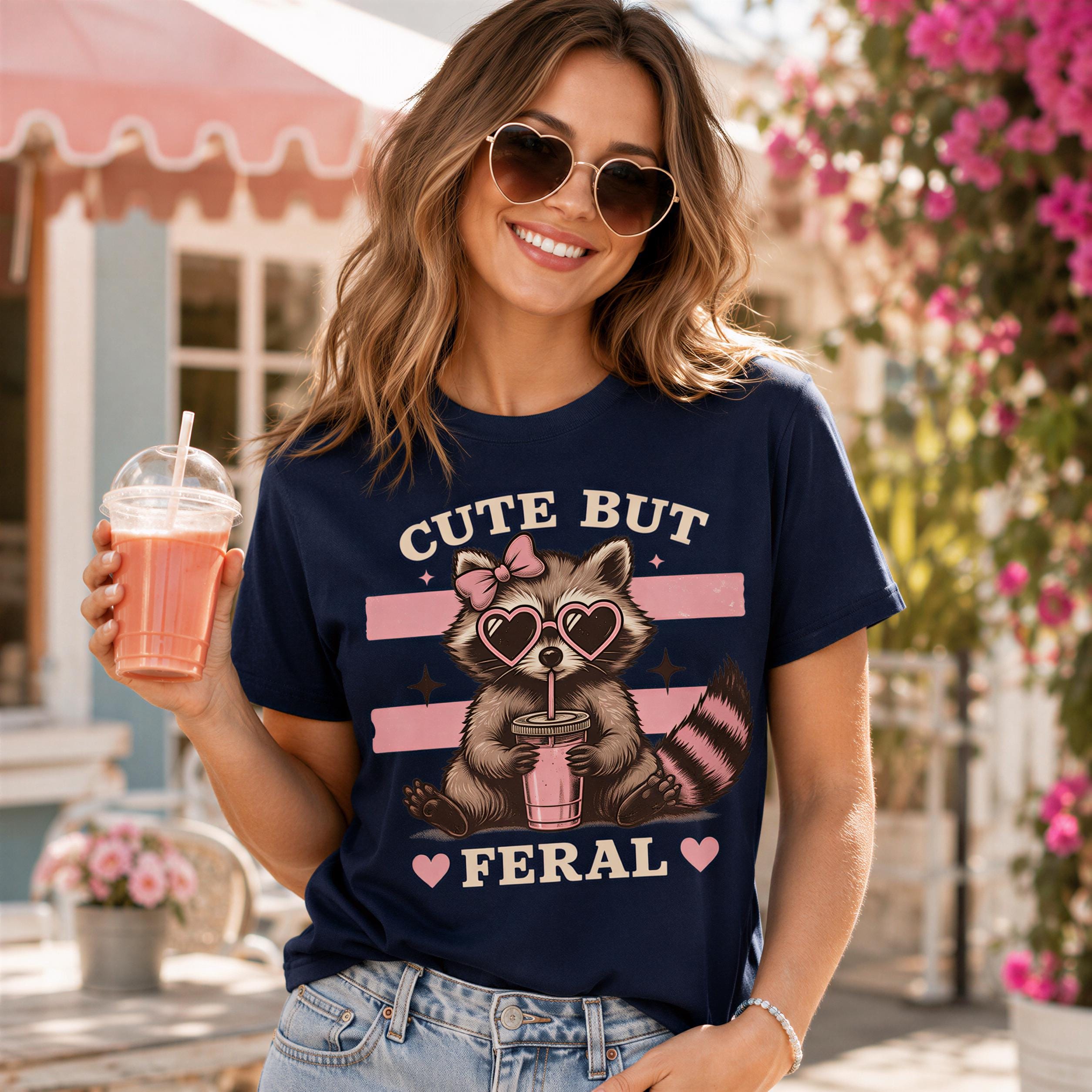 A woman wearing a navy blue t-shirt with a graphic of a raccoon and the text "CUTE BUT FERAL" is holding a drink and smiling.
