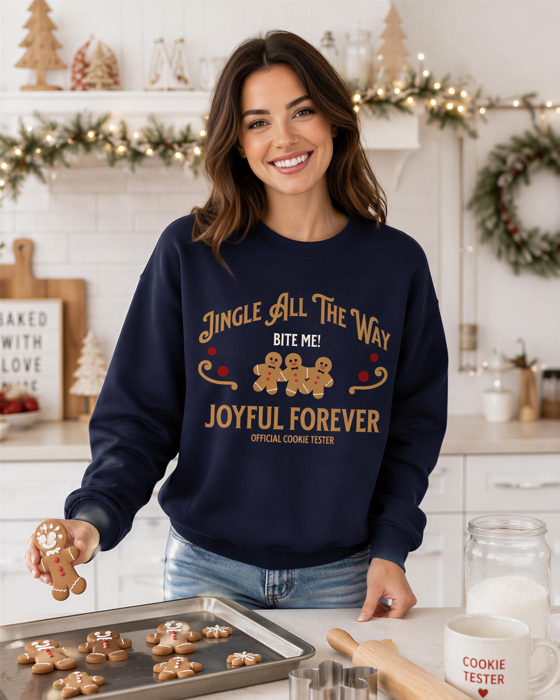 A woman is standing in a kitchen, smiling and holding a tray of freshly baked gingerbread cookies. She is wearing a navy blue sweatshirt with a humorous text that reads "Jingle all the way bite me! Joyful forever Official Cookie Tester".