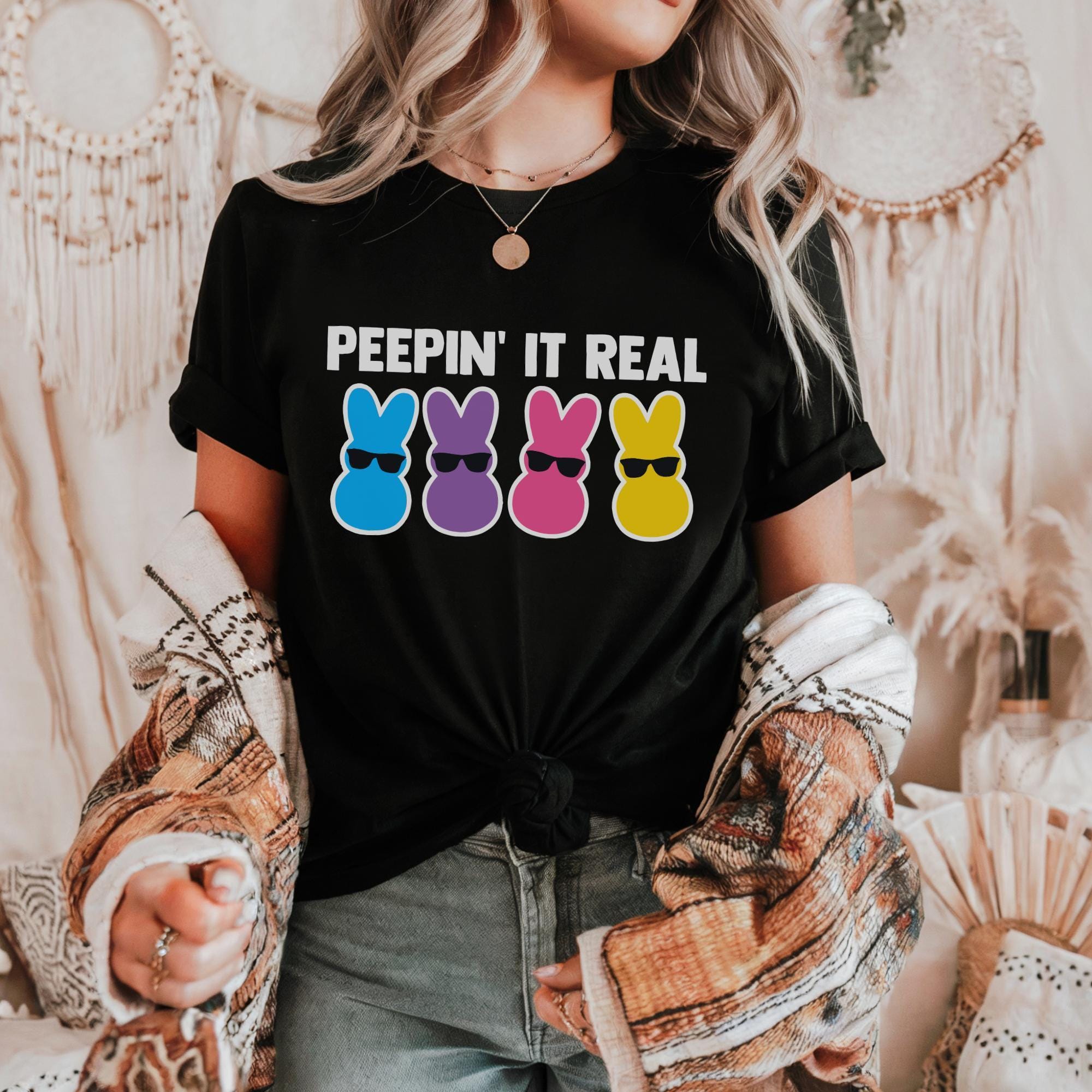 A woman wearing a black t-shirt with a graphic design of four colorful Easter bunny characters and the text "Peepin' It Real" printed on it.