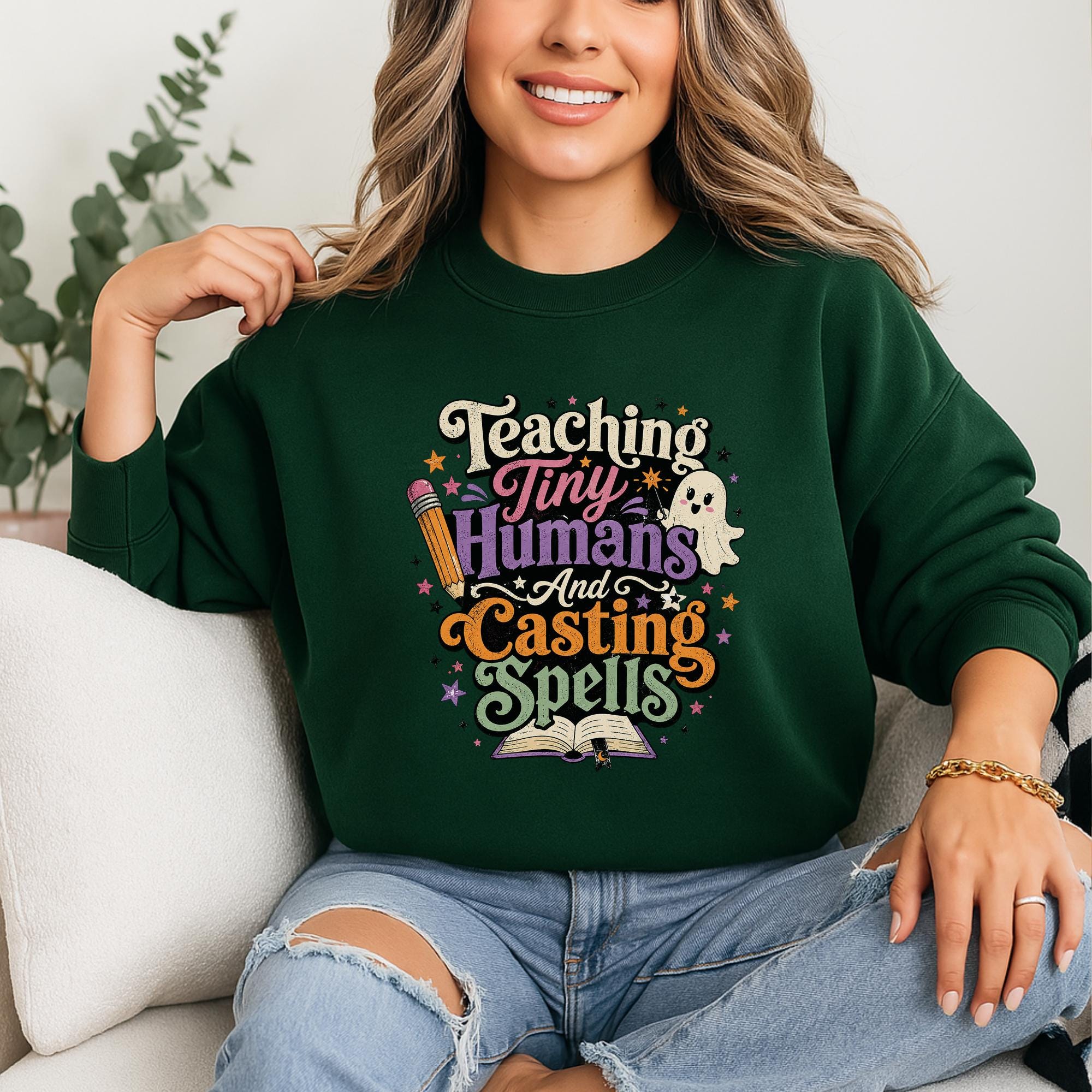 A woman wearing a green sweatshirt with a colorful graphic design that says "Teaching Tiny Humans and Casting Spells".