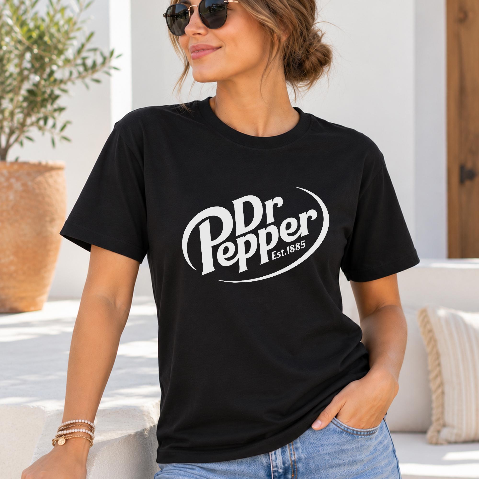 A woman wearing a black t-shirt with the Dr Pepper logo and jeans is standing in front of a potted plant.