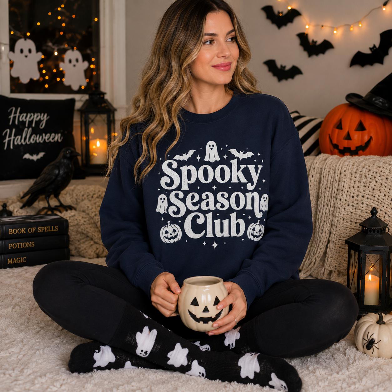A woman sitting on the floor wearing a navy blue sweatshirt with the text "Spooky Season Club" and holding a mug, surrounded by Halloween decorations and a pumpkin.